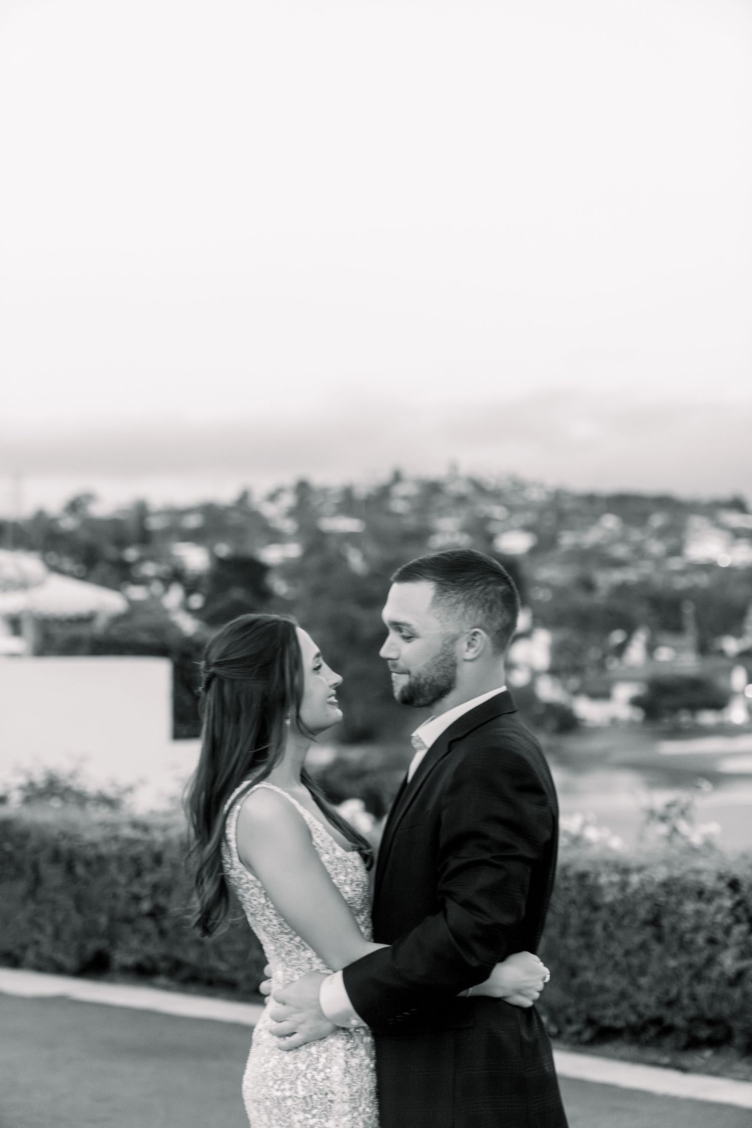 Intimate black and white portrait of couple in formal attire against a scenic hillside backdrop.
