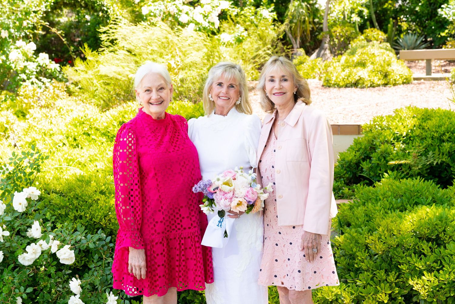 Three women in celebratory attire pose together in a lush garden setting with floral bouquet.