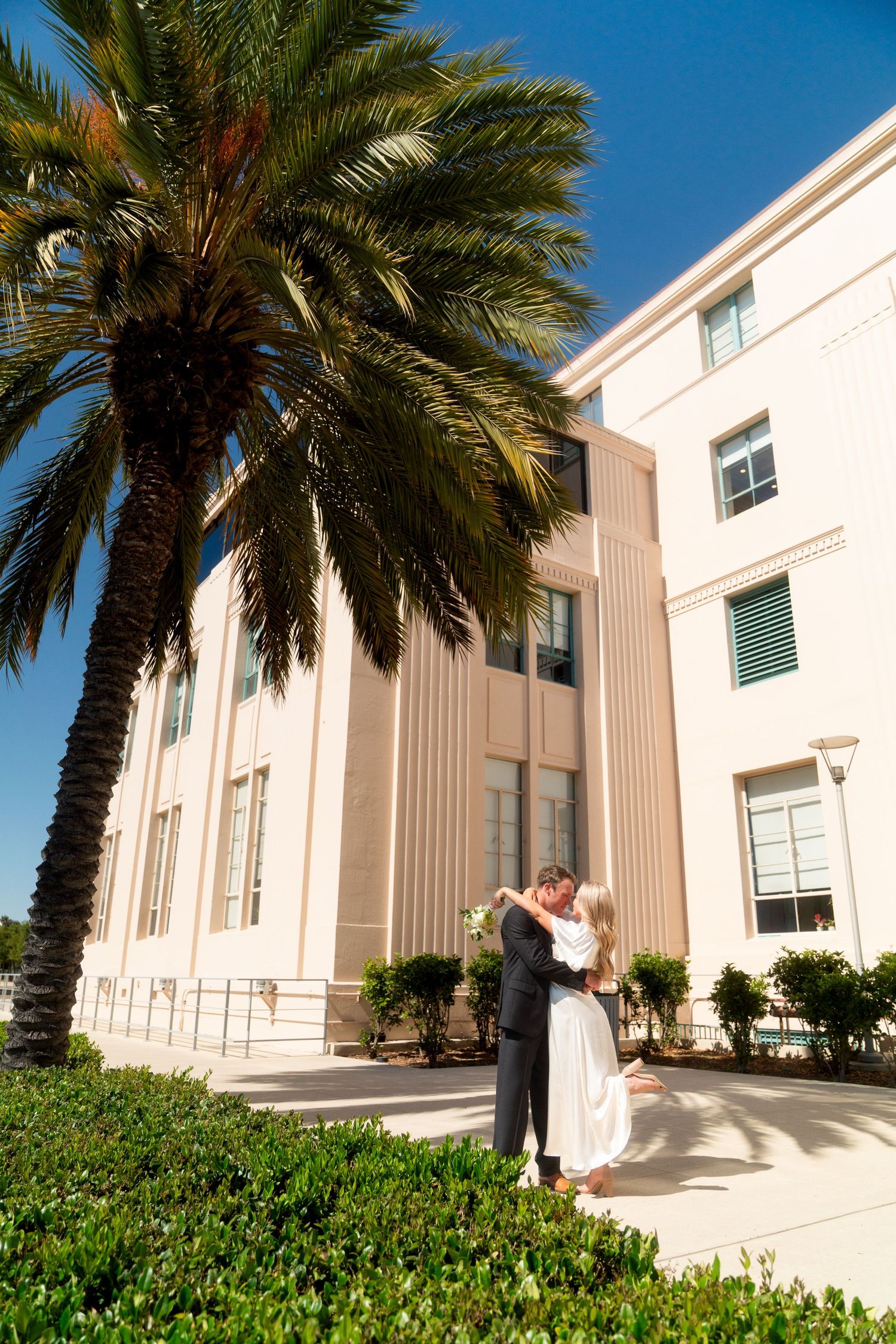 Modern white building exterior with palm trees and landscaping against bright blue sky on sunny day.
