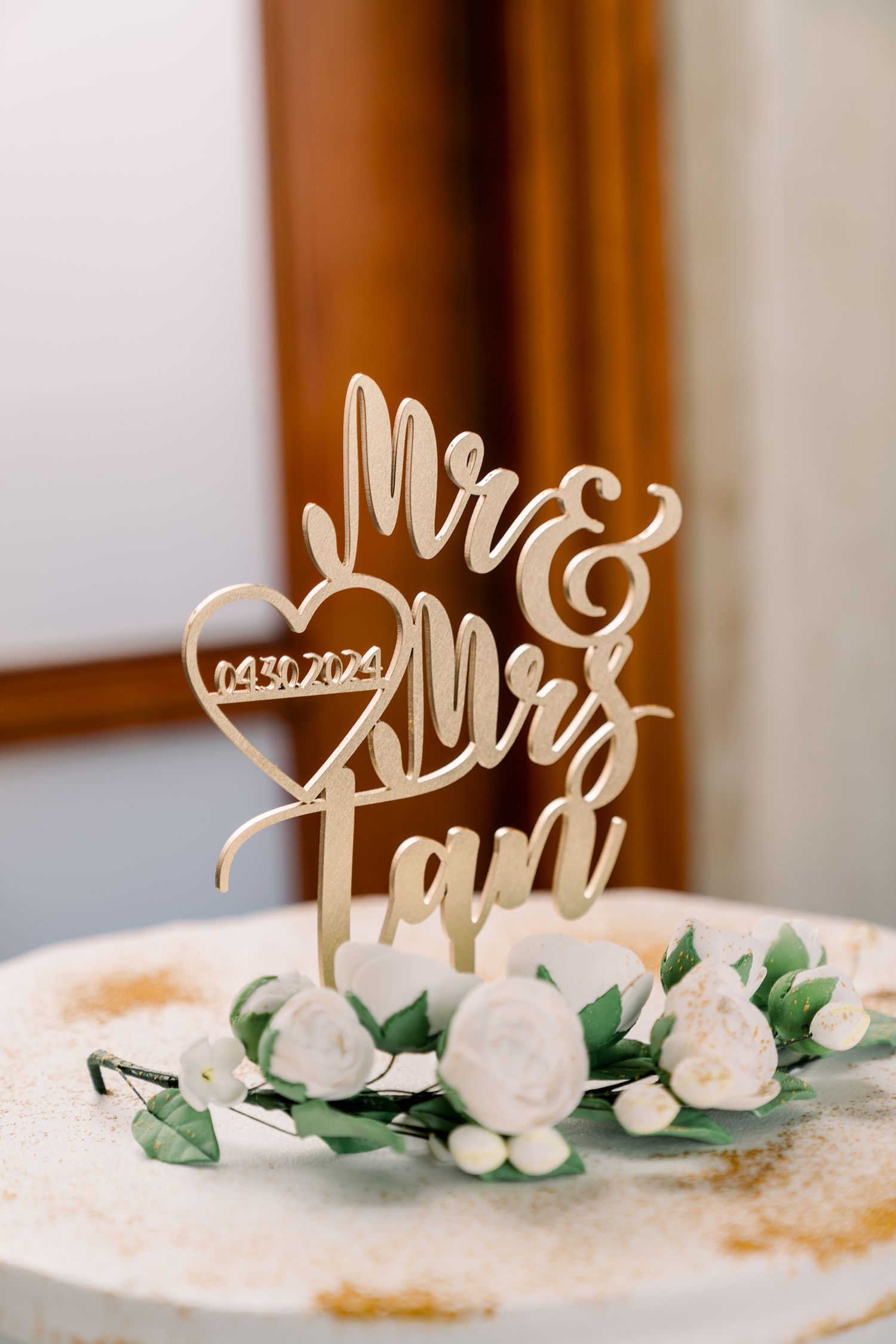 Wedding cake topper reading 'Mr & Mrs' with a heart date detail surrounded by white roses and greenery.