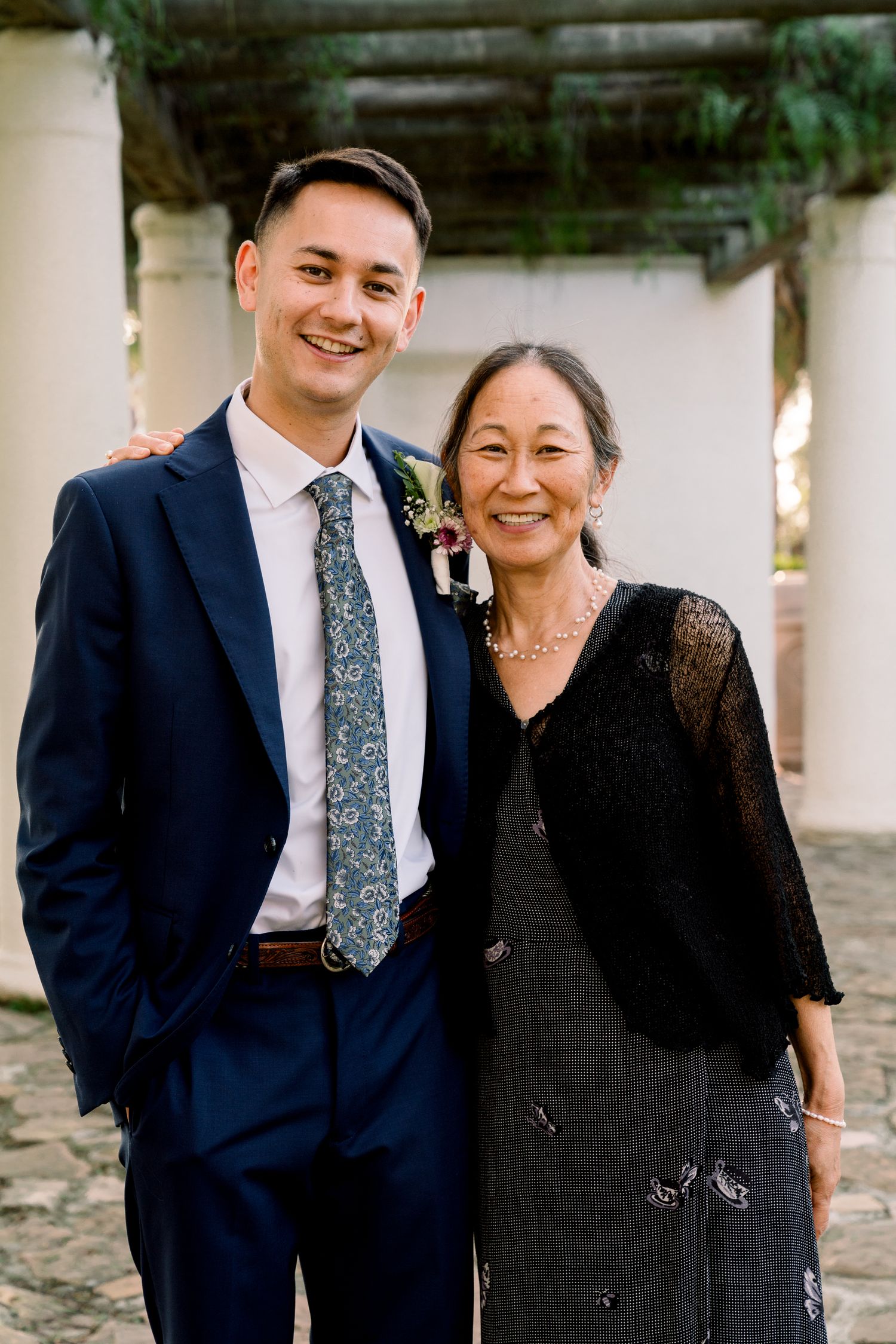 A well-dressed pair in formal attire stand together at what appears to be a wedding or special event venue.