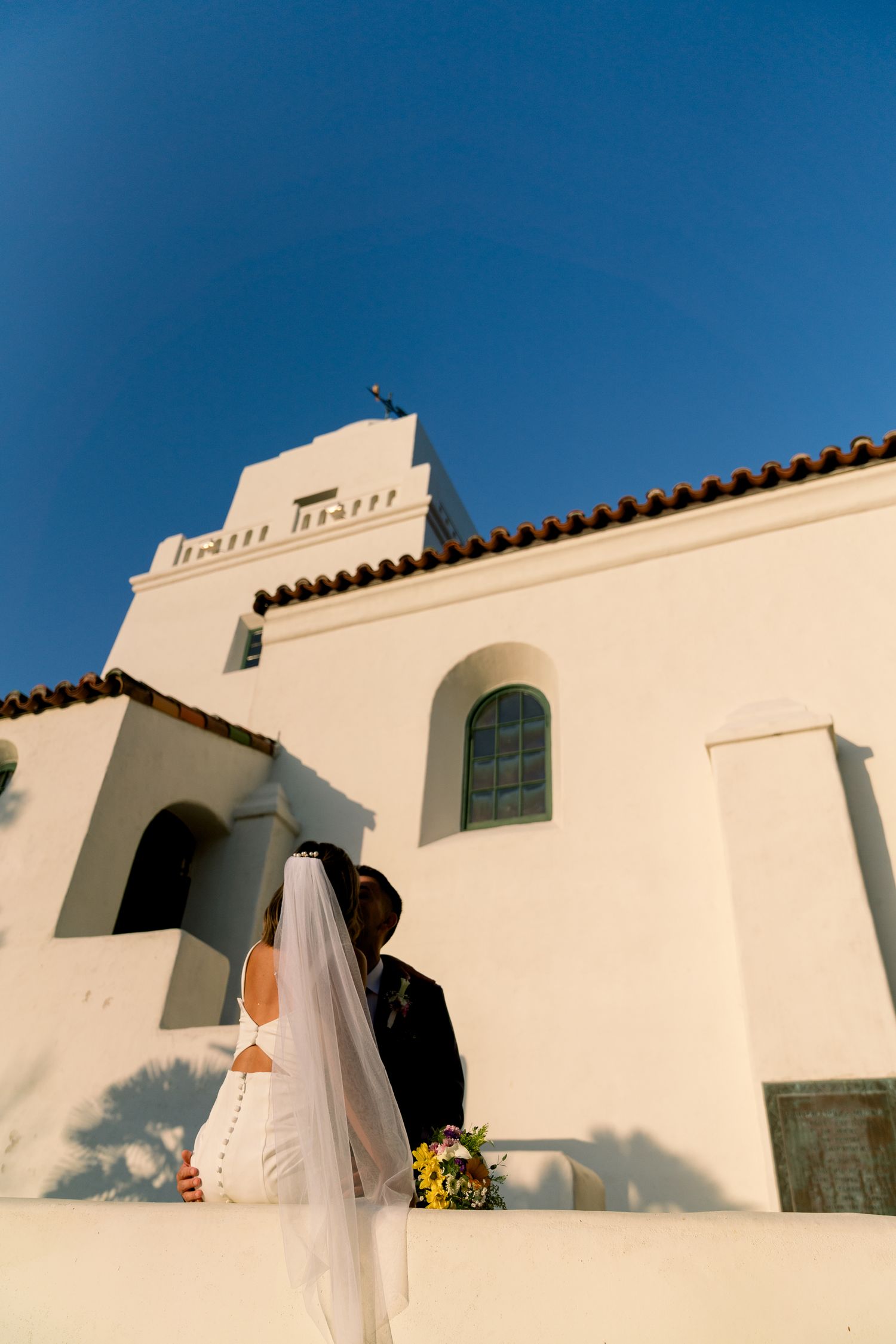 White Spanish mission-style church facade with terracotta roof tiles against deep blue sky.