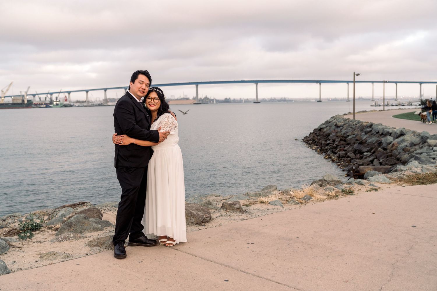 Couple embracing by the water with Coronado Bridge visible in background at sunset.