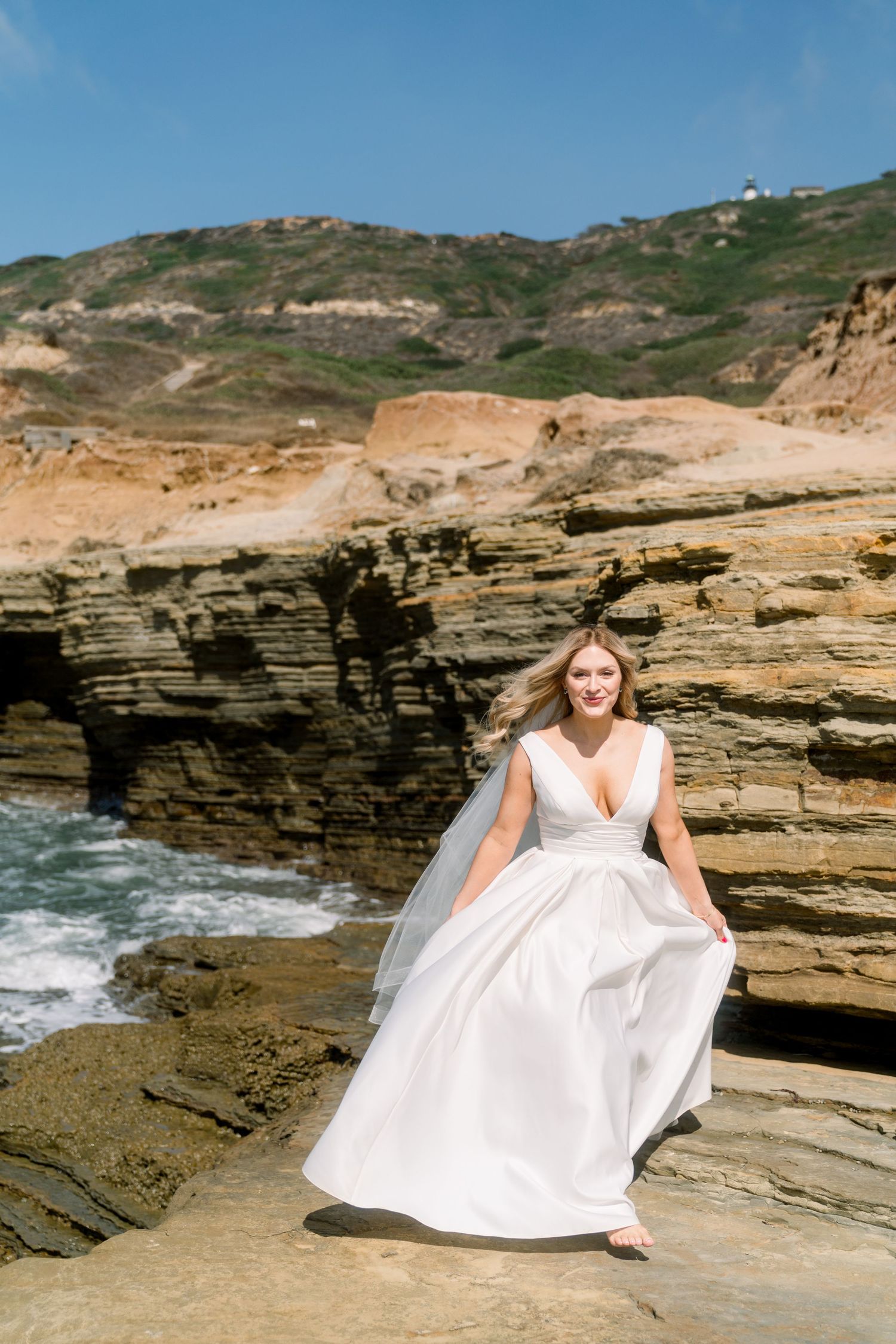 A wedding dress flows elegantly against dramatic coastal cliffs and crashing waves at a scenic oceanside location.