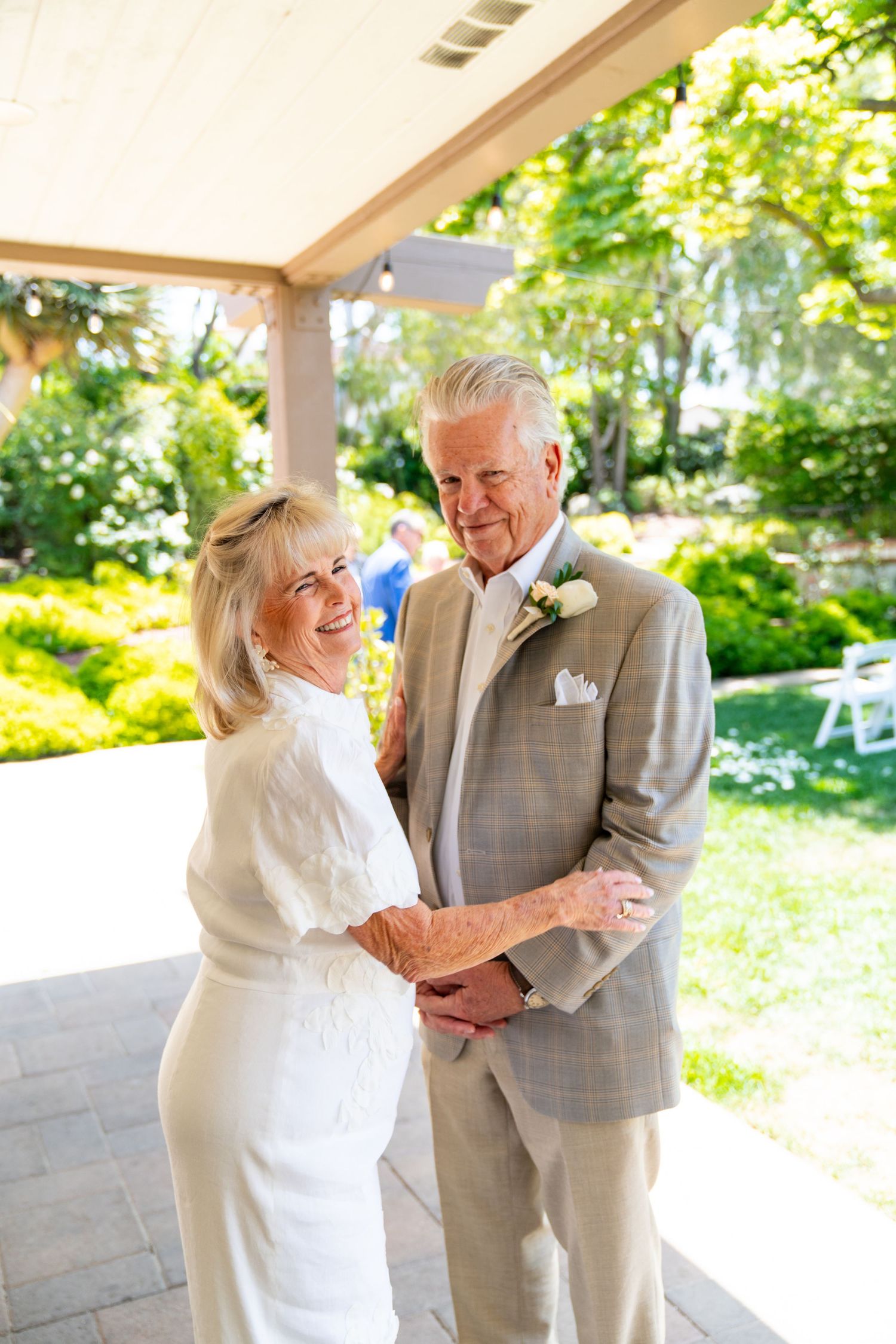 Senior couple in formal attire share a romantic moment dancing on a porch surrounded by lush garden scenery.