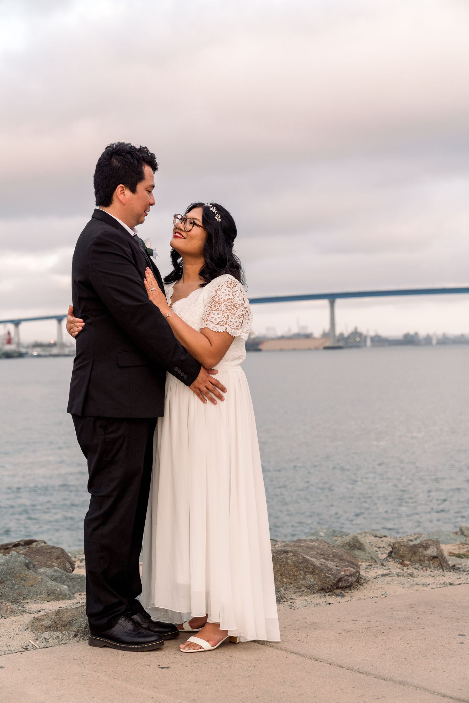 A couple in wedding attire stands on a rocky beach shore with the San Francisco Bay and bridge in the distance.