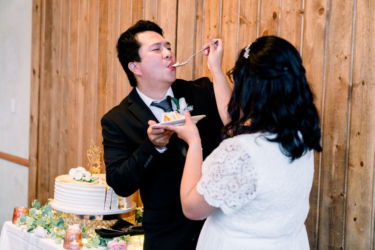 A couple shares their wedding cake during a reception in front of a rustic wooden wall.