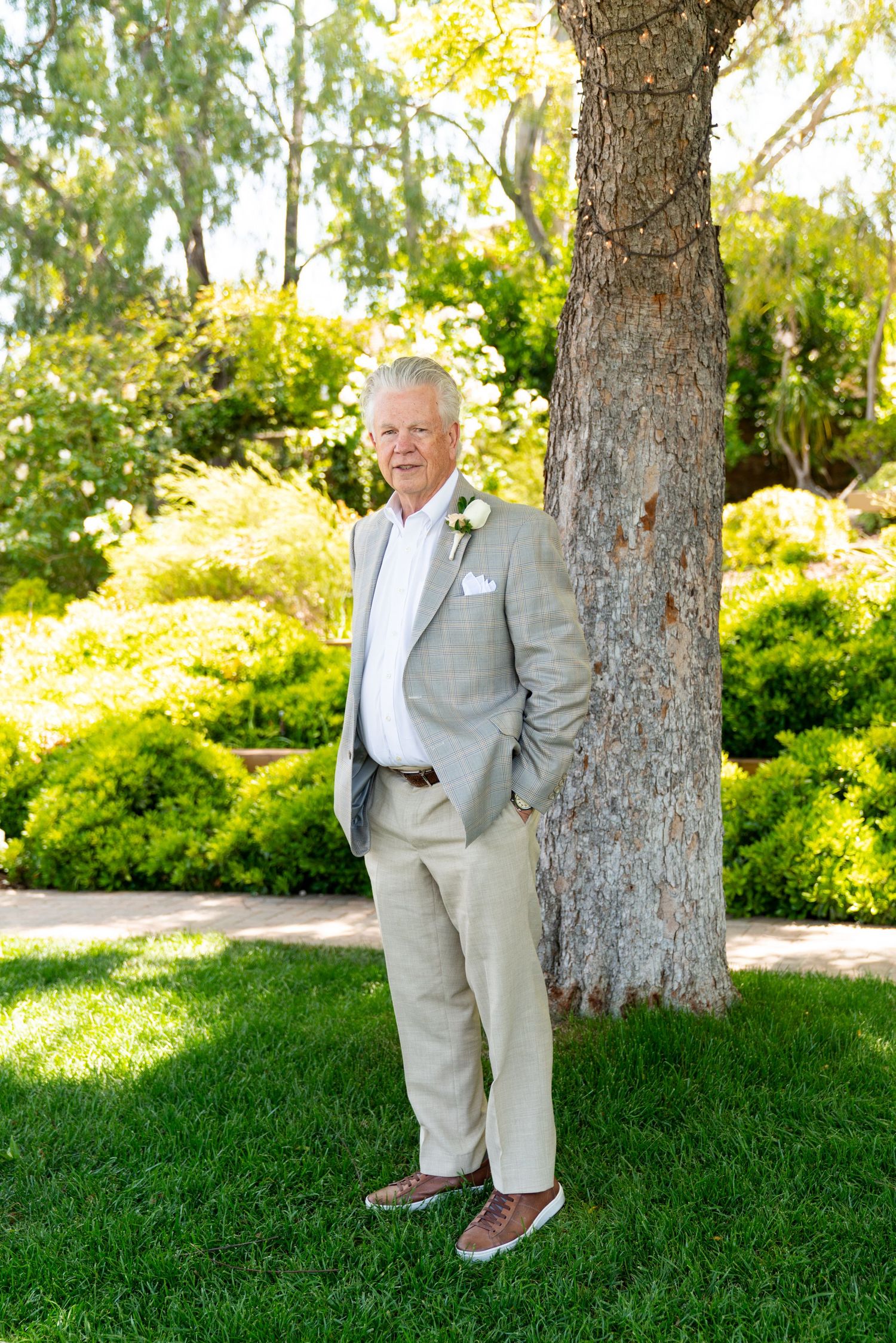 Person in a light gray suit and boutonniere stands casually next to a tree trunk in a sunny garden setting.