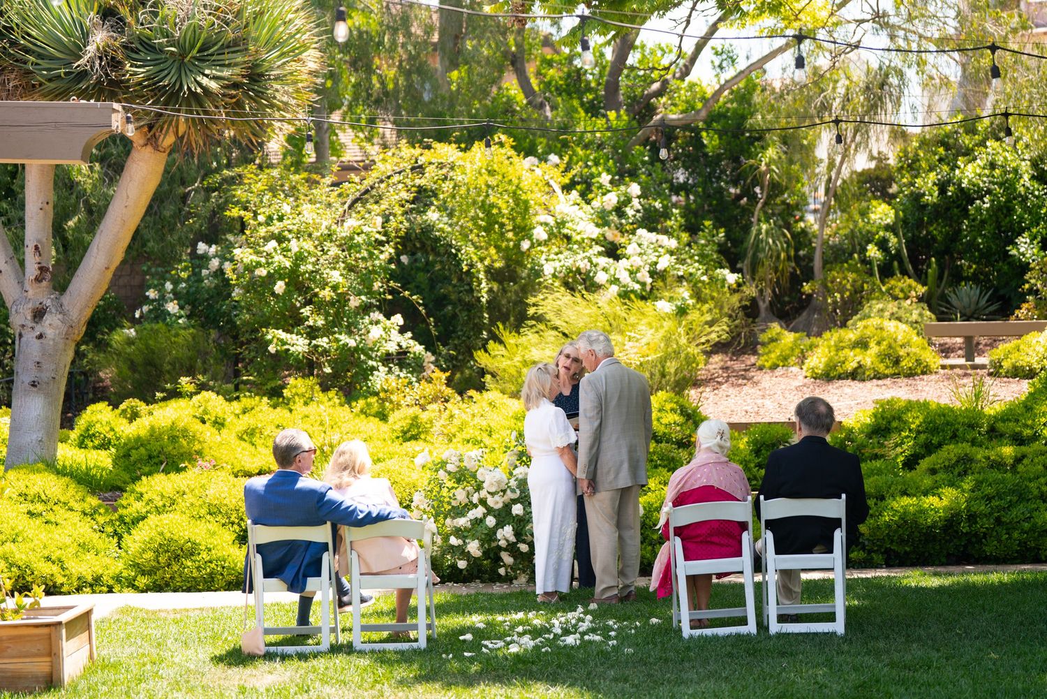 Small outdoor ceremony taking place in a lush garden setting with white chairs and vibrant yellow florals.