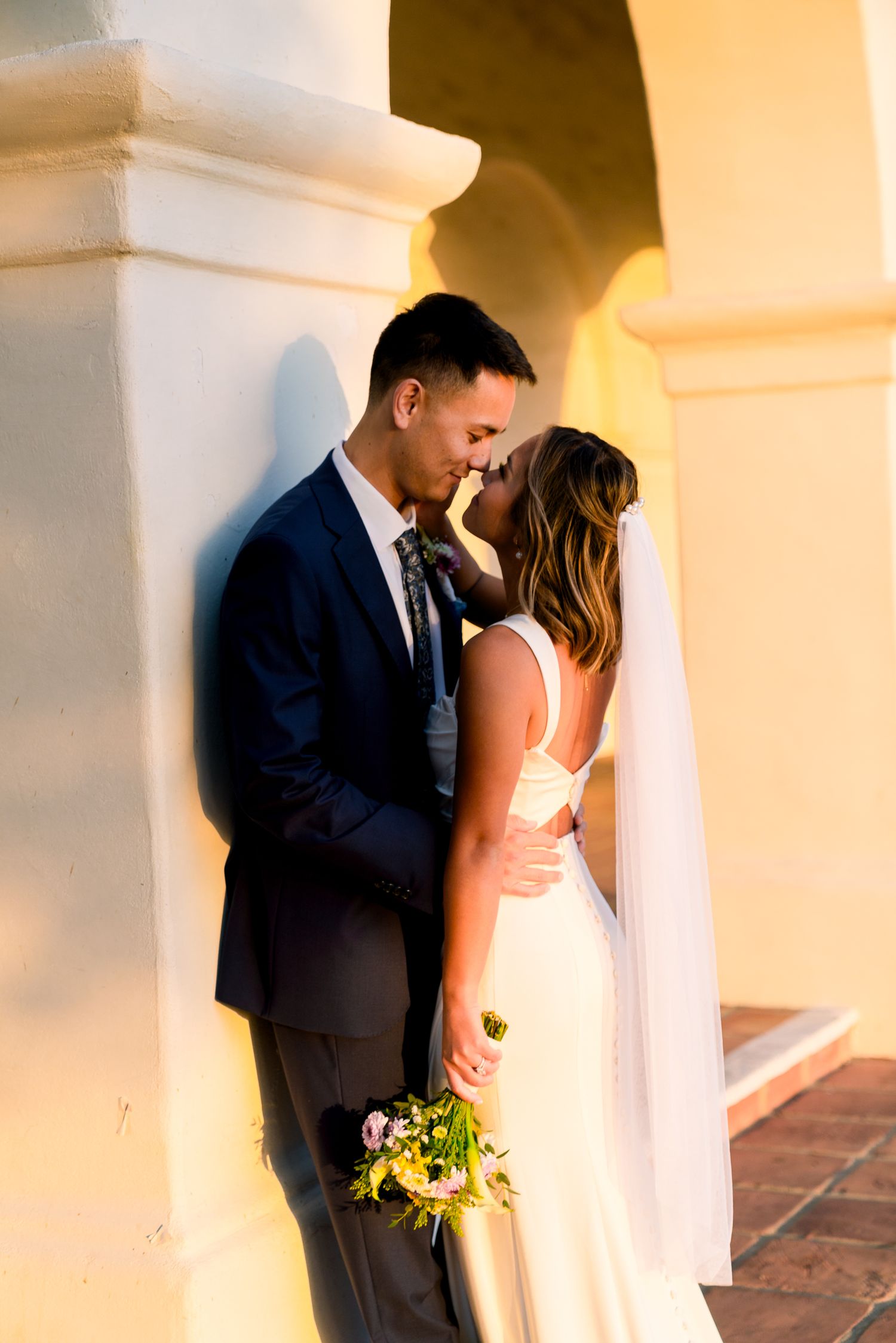 Intimate wedding portrait with couple sharing tender moment against white column at sunset.