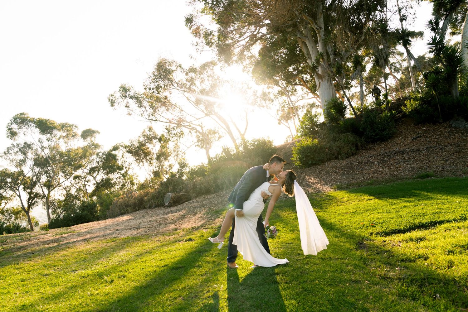 A couple shares a romantic moment on a grassy hillside at sunset with sunlight streaming through trees in the background.