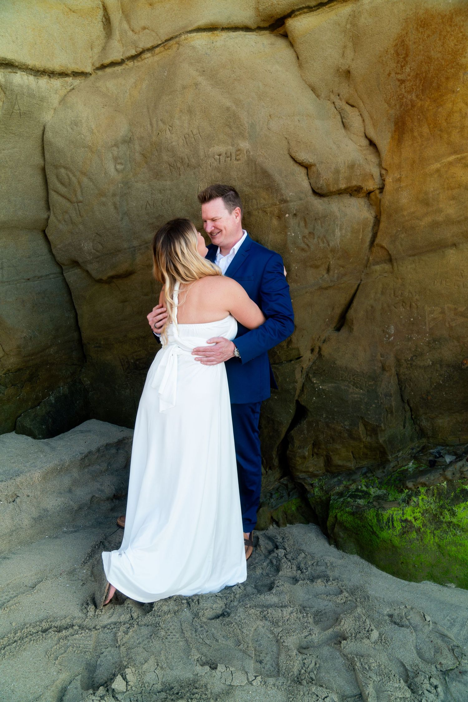 A couple in wedding attire embrace against dramatic rocky cliffs at what appears to be a beach or coastal setting.
