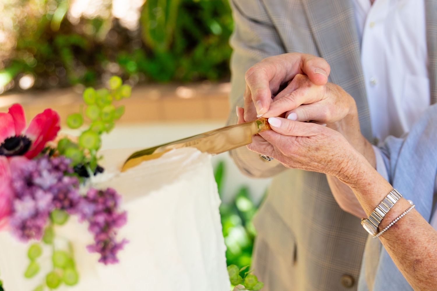 Hands working with art supplies and flowers at a table with colorful lilac and red blooms nearby.