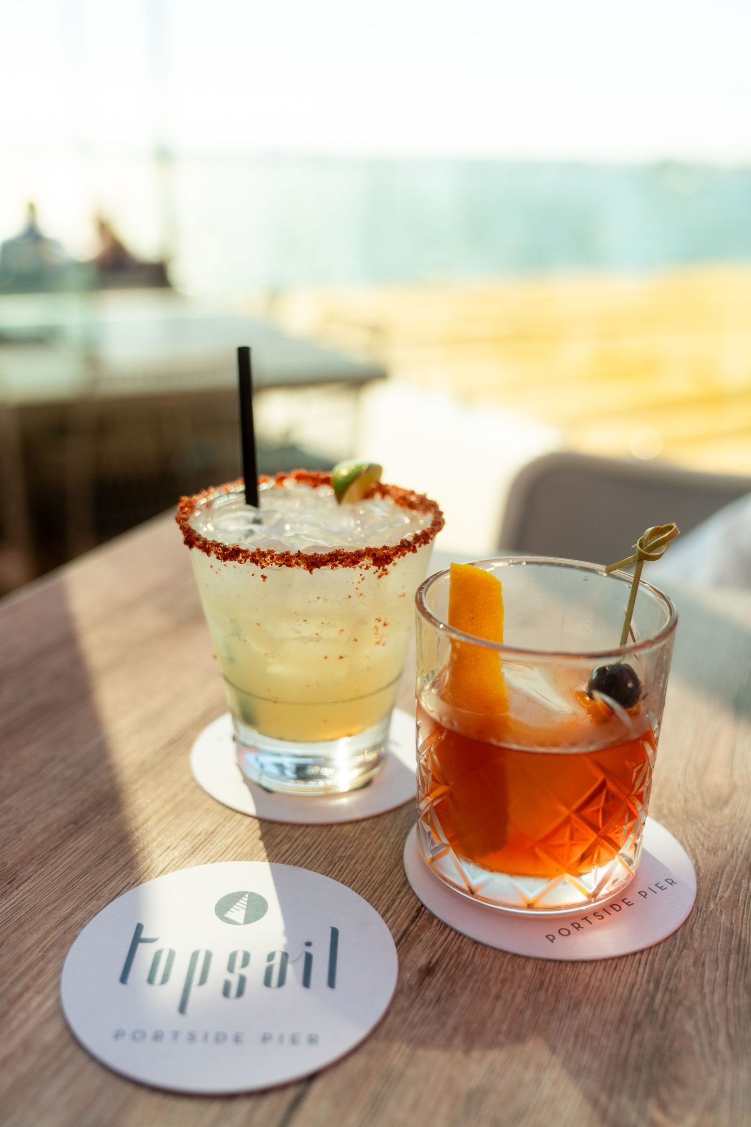 Two craft cocktails with garnishes sit on logo coasters at a bar, one margarita-style and one old fashioned.
