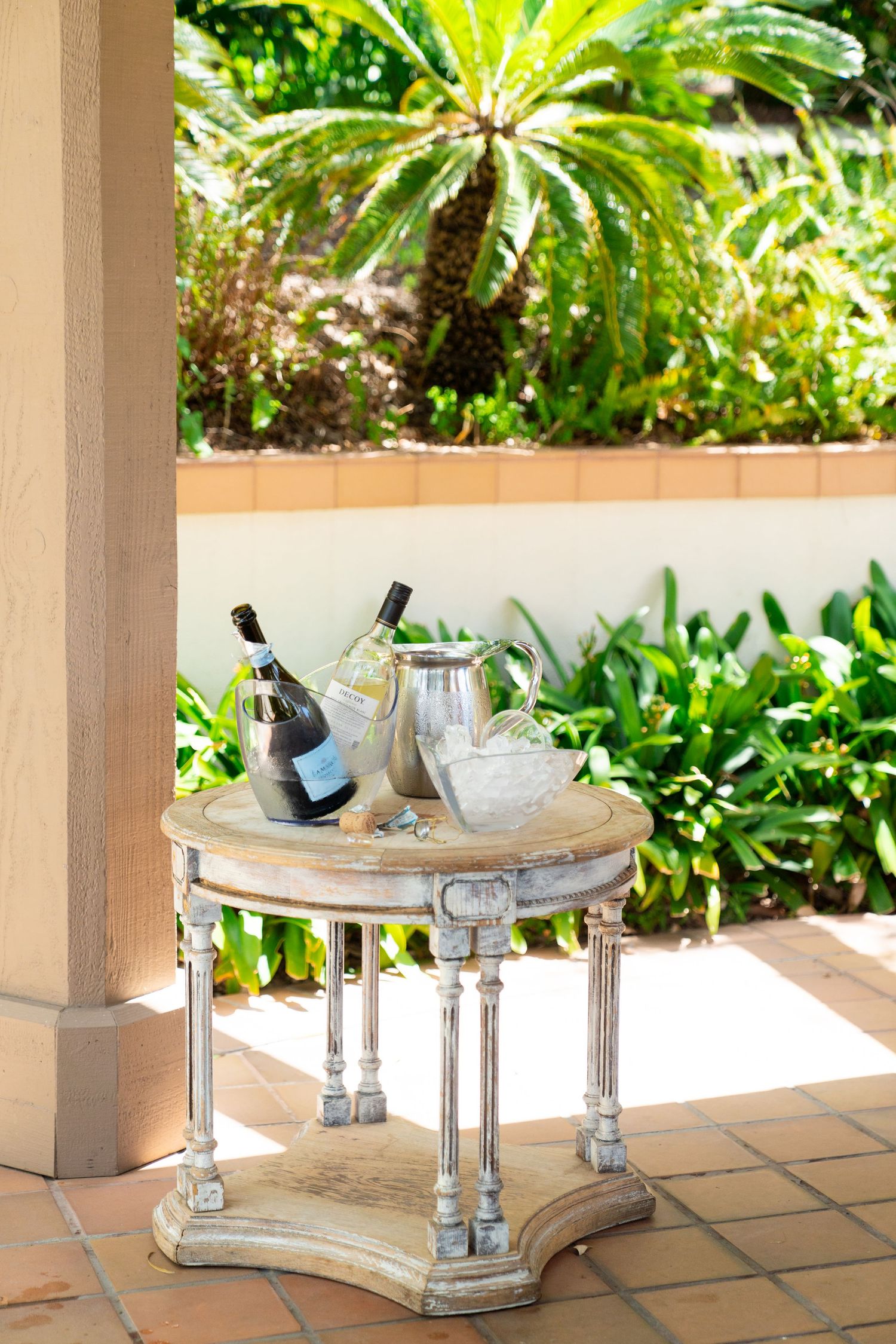 A rustic round side table with wine and glassware sits on a tiled patio surrounded by tropical plants.