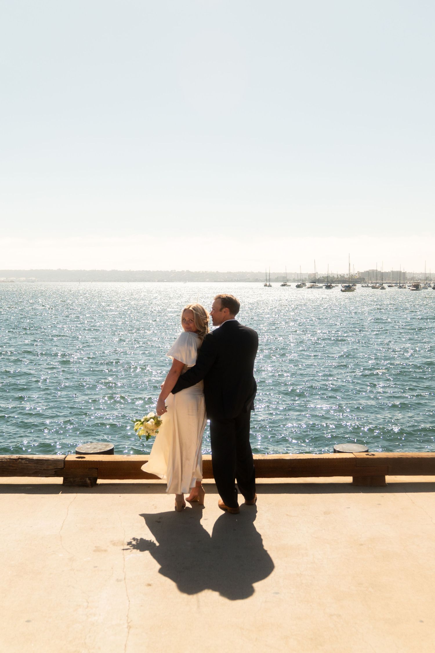 A couple in formal attire embraces while standing on a waterfront deck overlooking a harbor with boats on a sunny day.