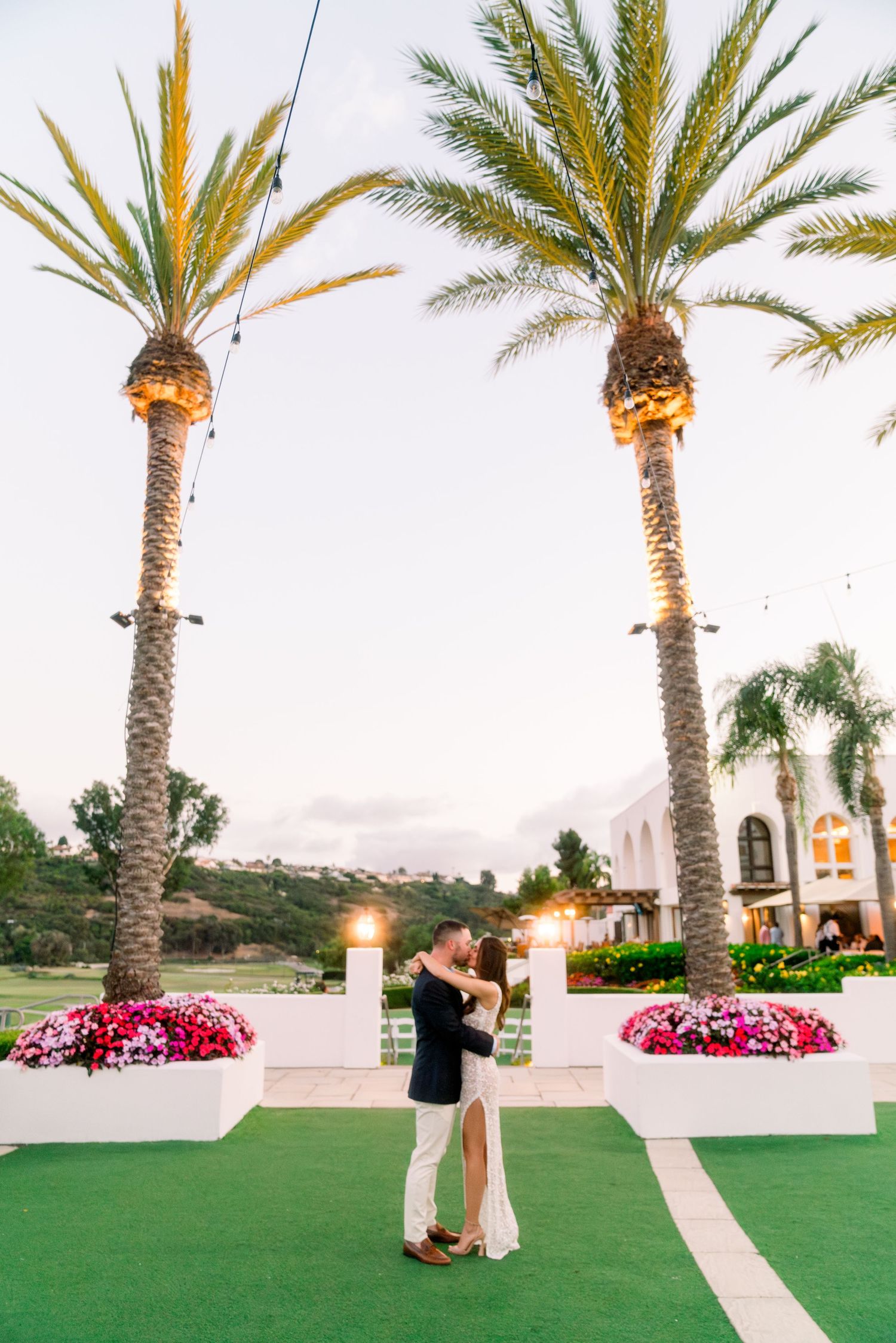 Couple dancing on green terrace between two tall palm trees with pink flowers in planters.
