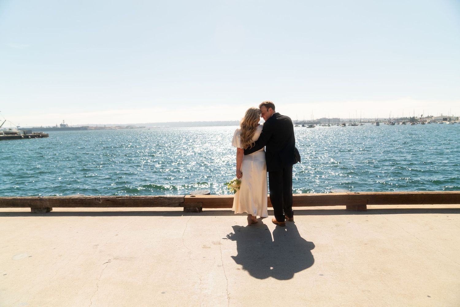 A couple stands together on a sunny waterfront pier overlooking a beautiful harbor on their wedding day.