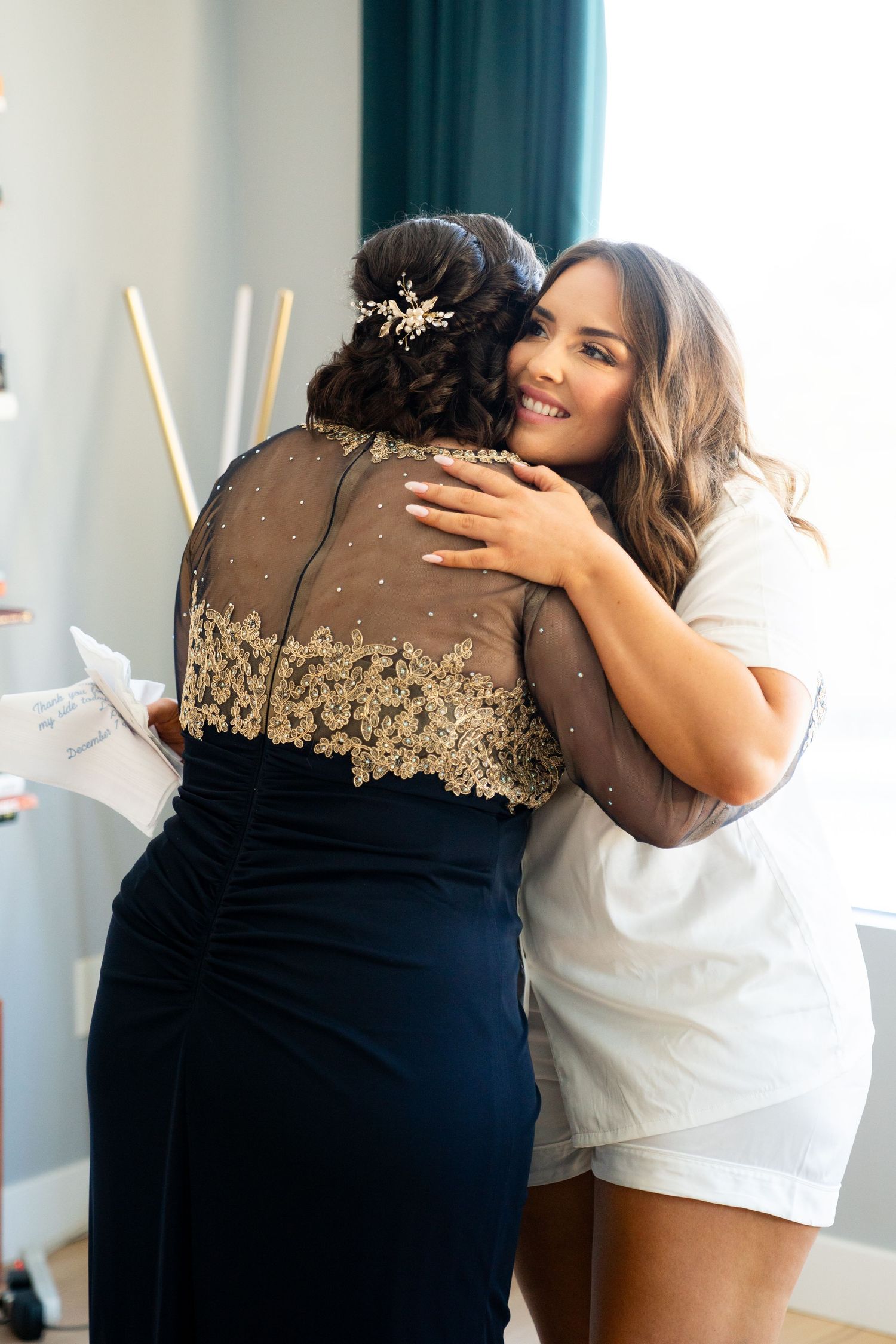 Two women share an emotional embrace, one wearing a navy dress with gold lace detail and the other in white.