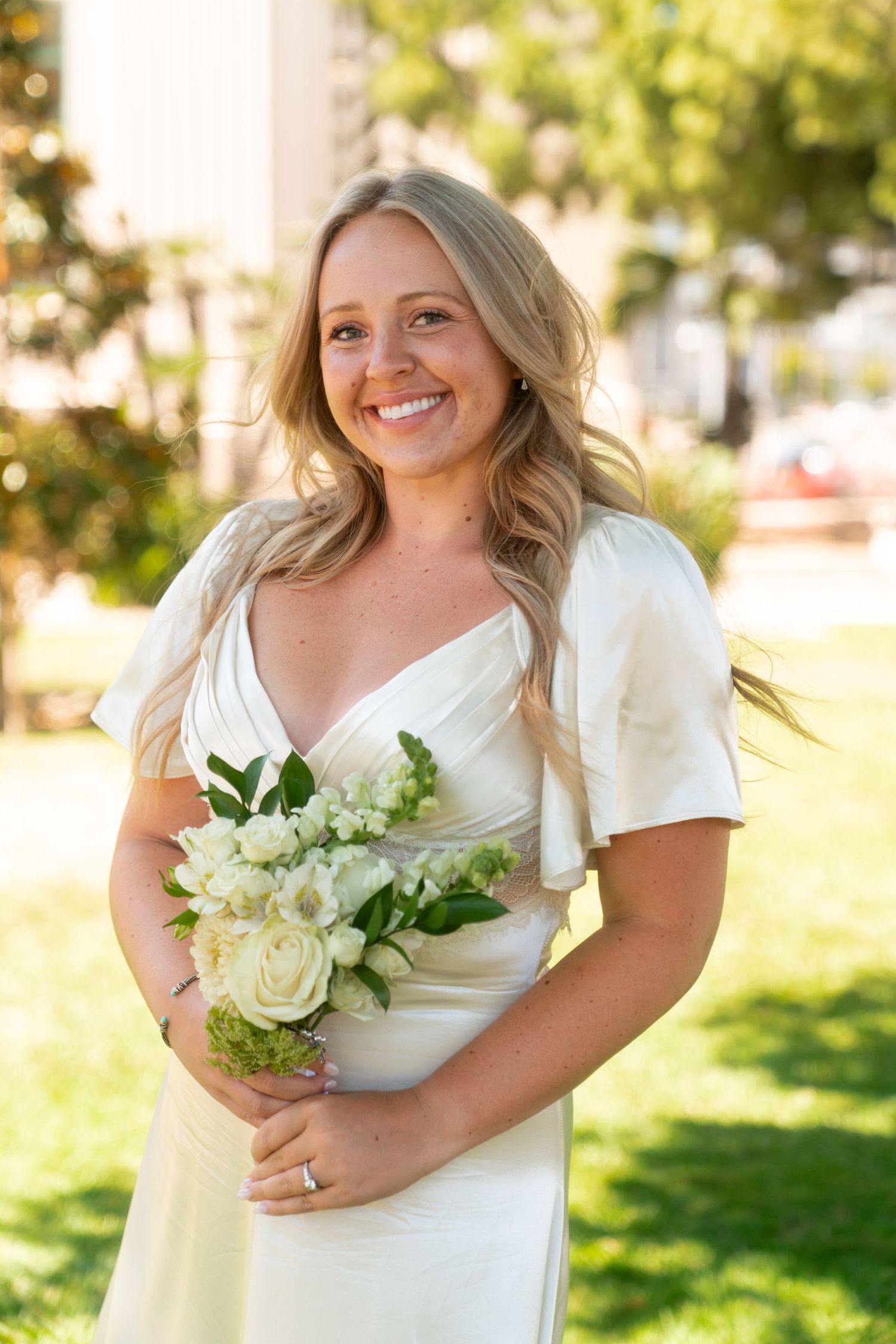 A bride in white dress with short flutter sleeves holds bouquet of cream roses in sunny outdoor setting.