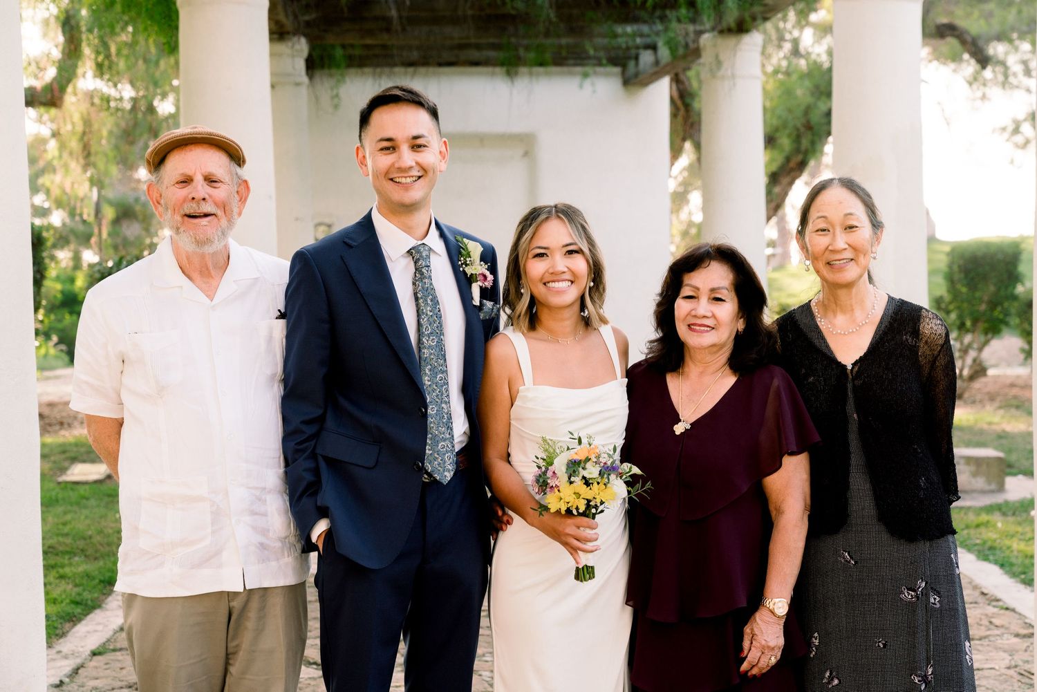 A happy group of five people standing together outdoors under a covered walkway, with one holding a bouquet of flowers.