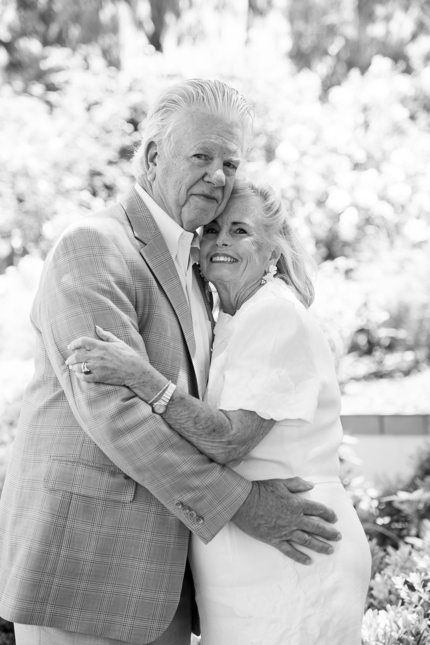An elderly couple embraces in a tender moment, sharing a joyful hug outdoors in black and white photography.