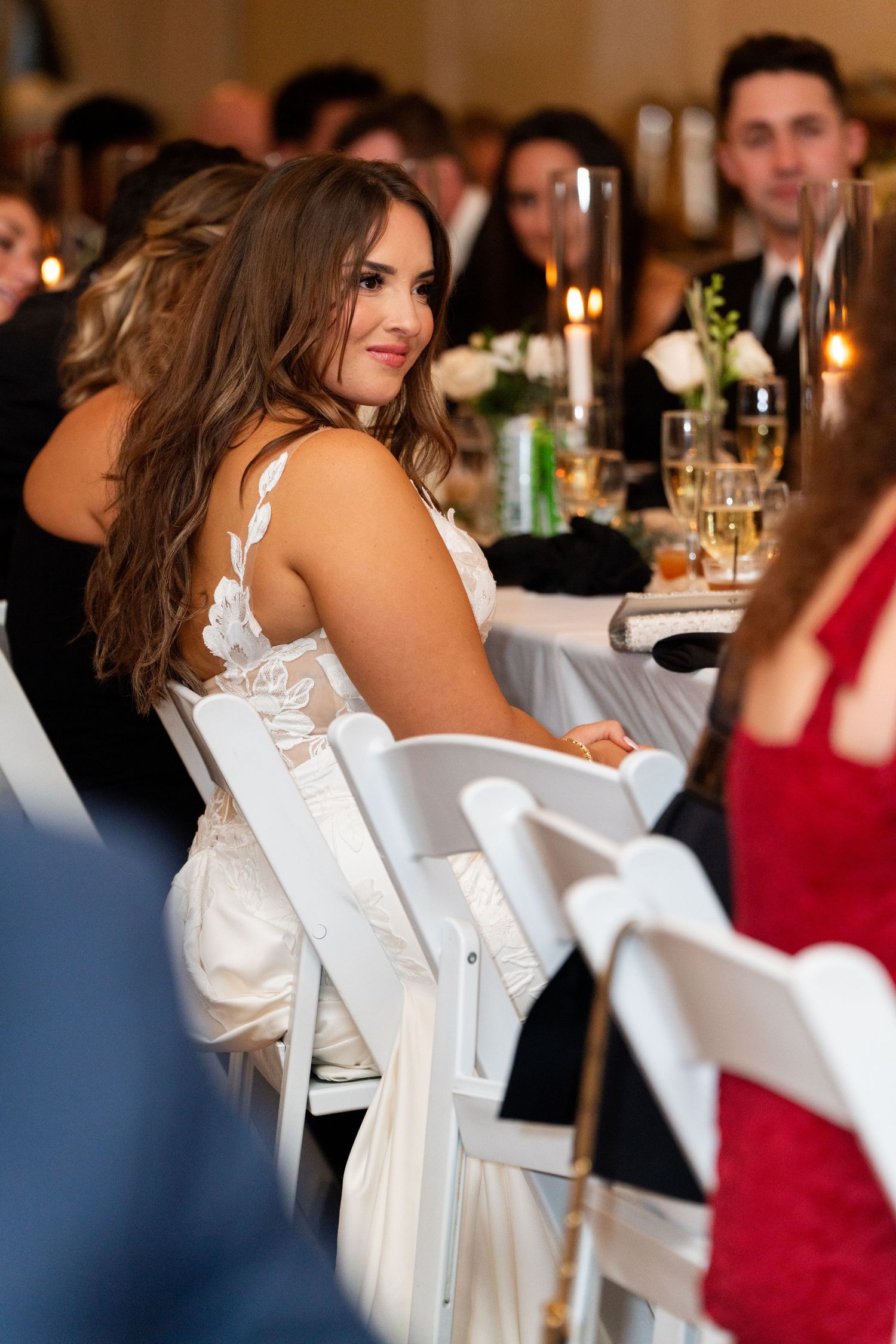 A bride in a white lace gown turns to smile during a wedding reception with candlelit tables in the background.
