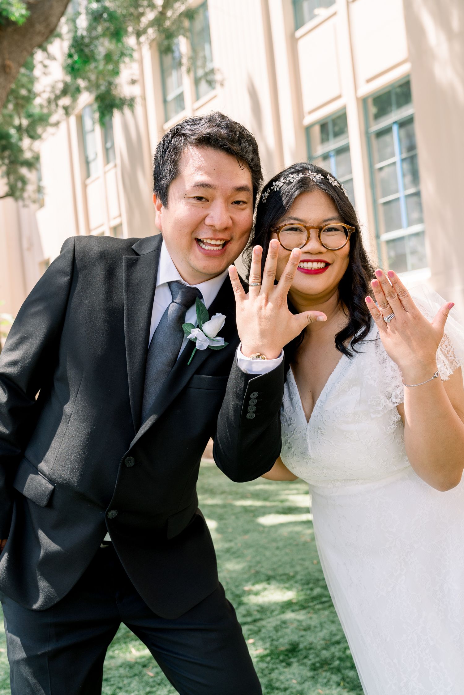 Newlyweds showing off their rings with joyful expressions outside a building.