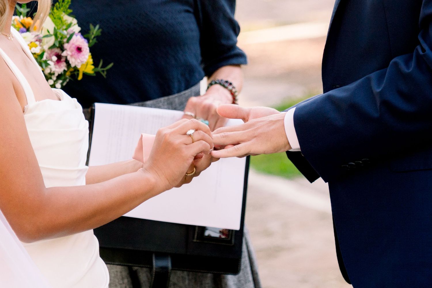 Close-up photo of a couple exchanging wedding rings during an outdoor ceremony with flowers in the background.
