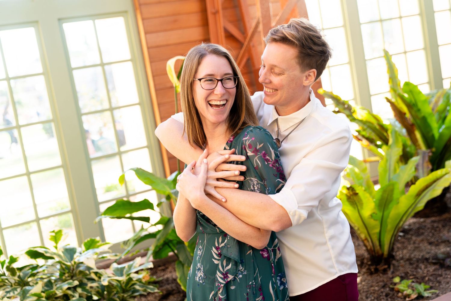 A joyful couple laughs together in a bright sunlit room with large windows and indoor plants.