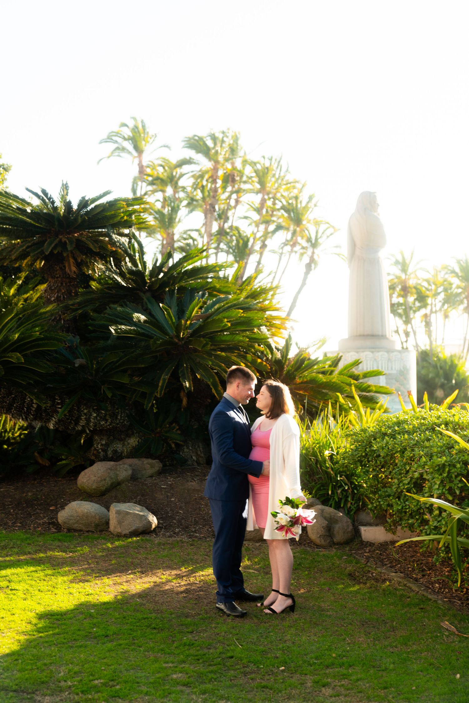 A couple stands together on a grassy lawn surrounded by palm trees and tropical foliage with a statue in the distance.