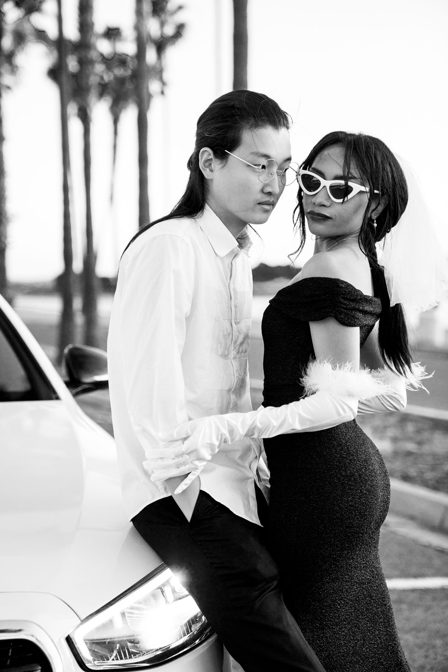 Stylish couple in formal attire posing against a car with palm trees in the background.