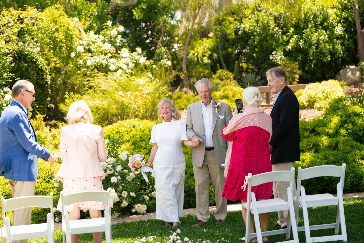 Guests gathering at outdoor garden ceremony with white chairs and lush greenery in background.