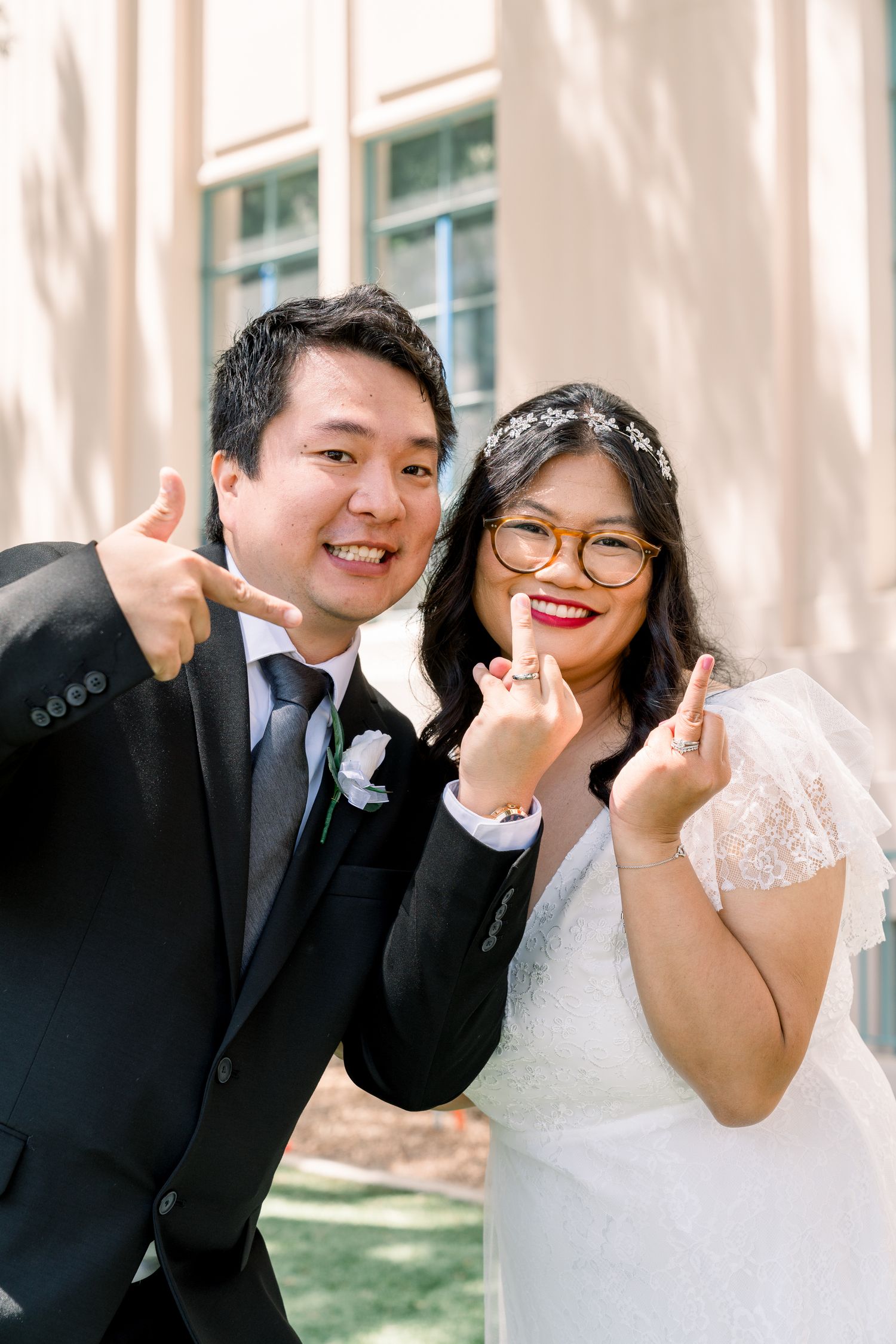 Wedding couple in formal attire making playful gestures while standing together outdoors.
