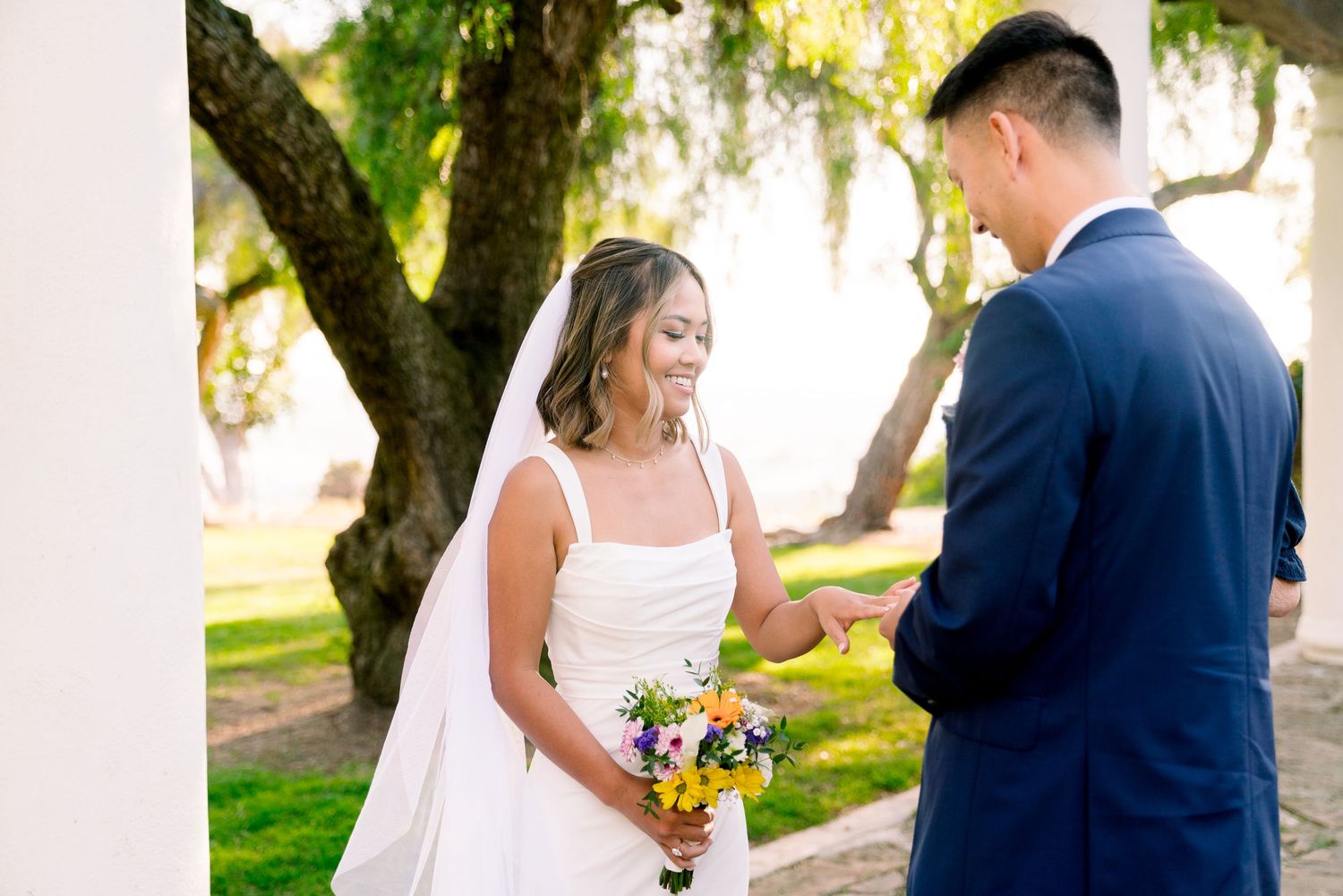Two people in wedding attire share a joyful moment together outdoors under a tree with sunlit foliage.