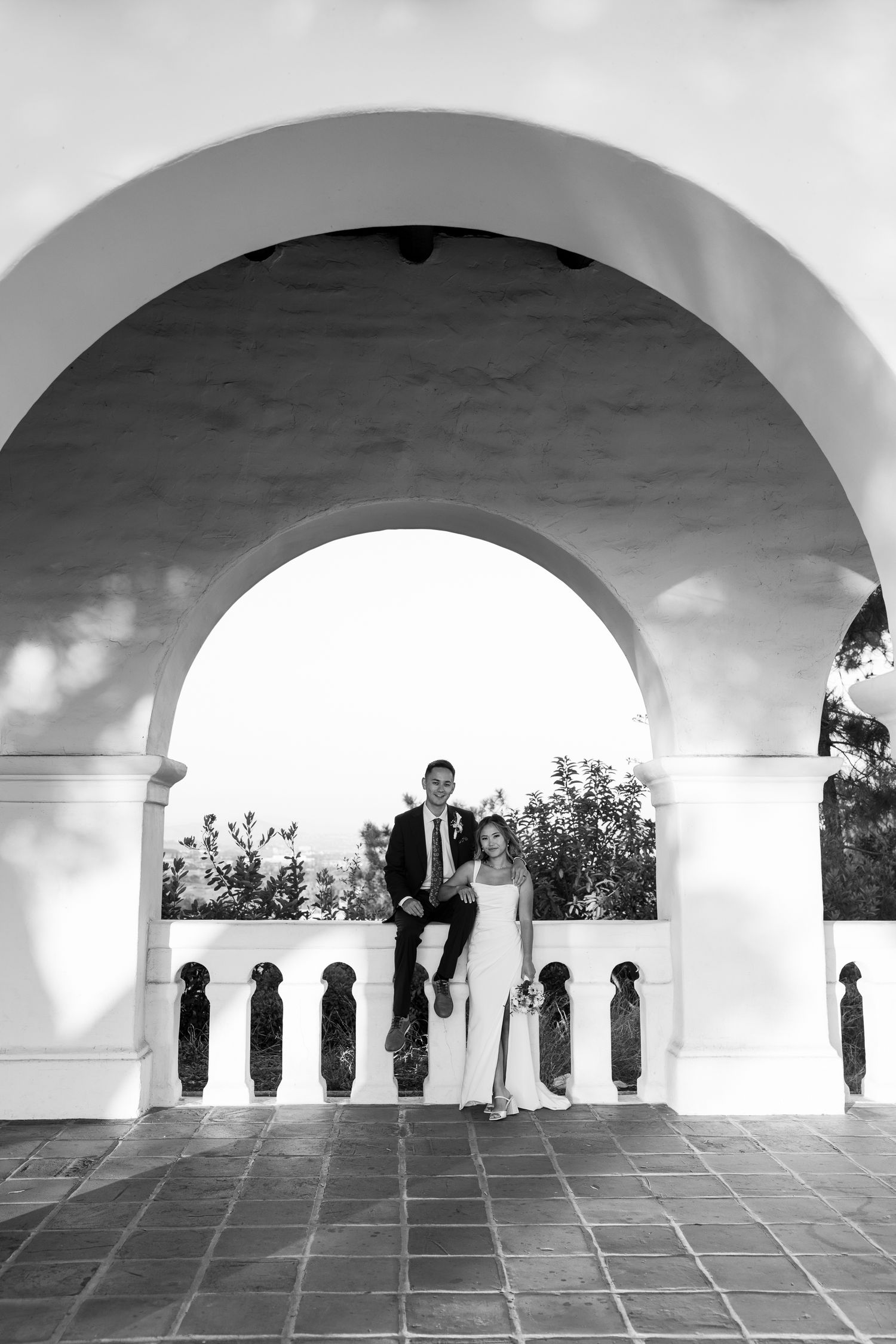 A couple sitting on white arched balcony railing under Spanish colonial architecture in black and white photography.