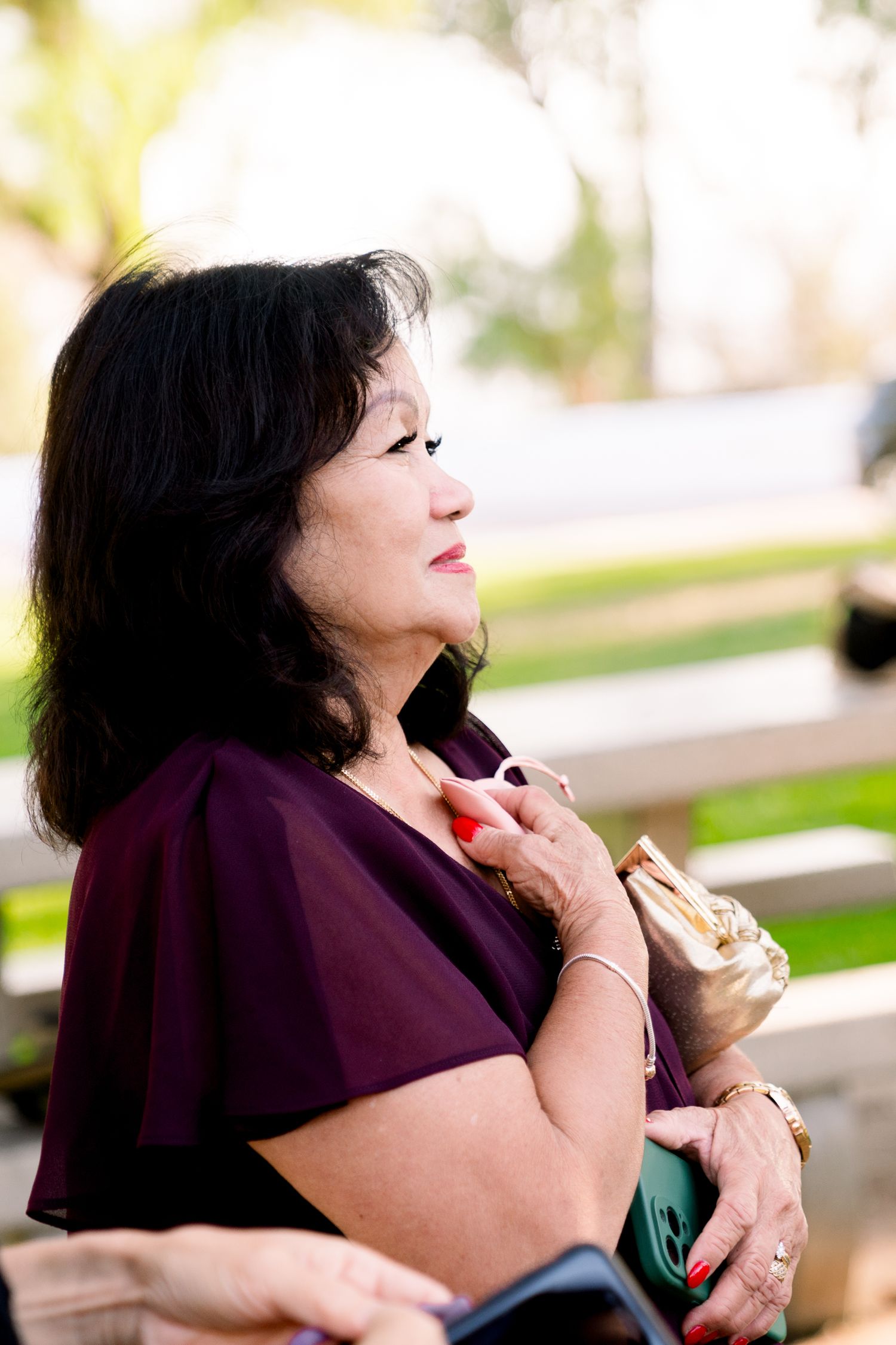 A woman in a purple blouse enjoys a beverage outdoors with soft sunlit bokeh in the background.