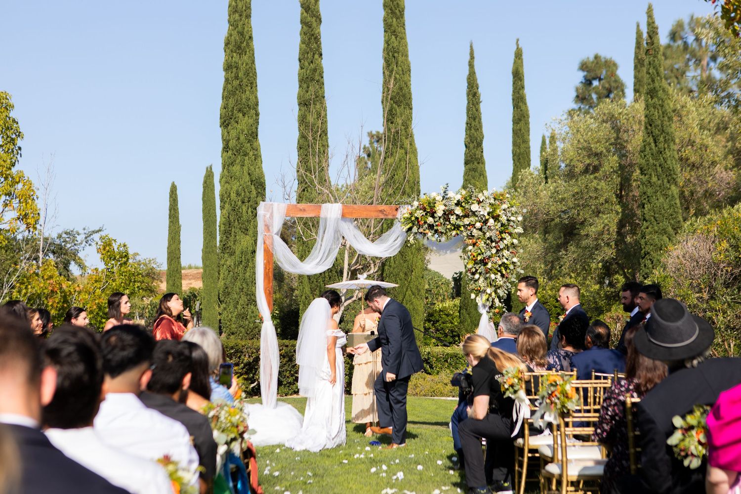 Outdoor wedding ceremony setup with cypress trees and floral arch in garden setting.