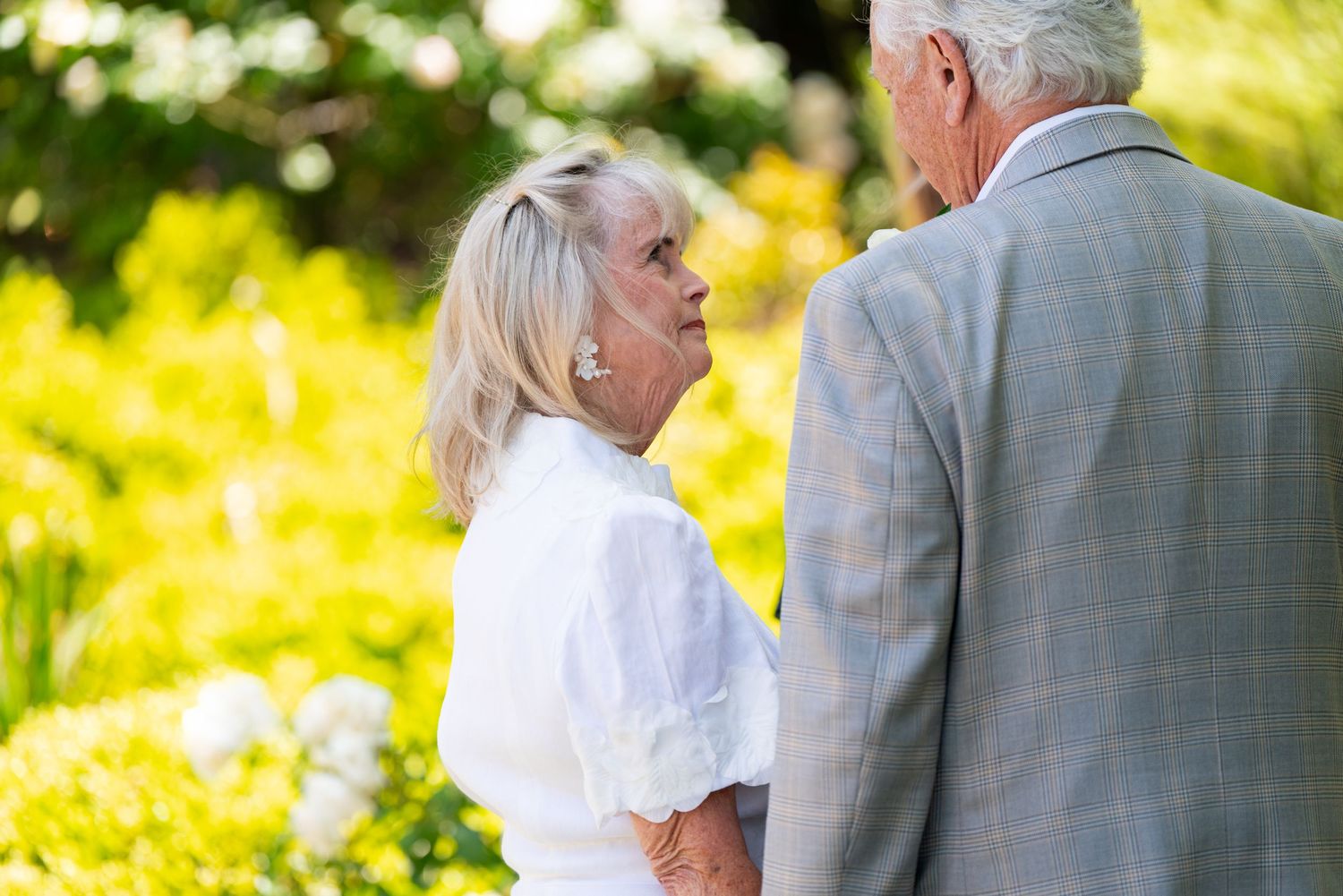 Two mature people share a loving moment in a sunny garden with yellow flowers blooming in the background.