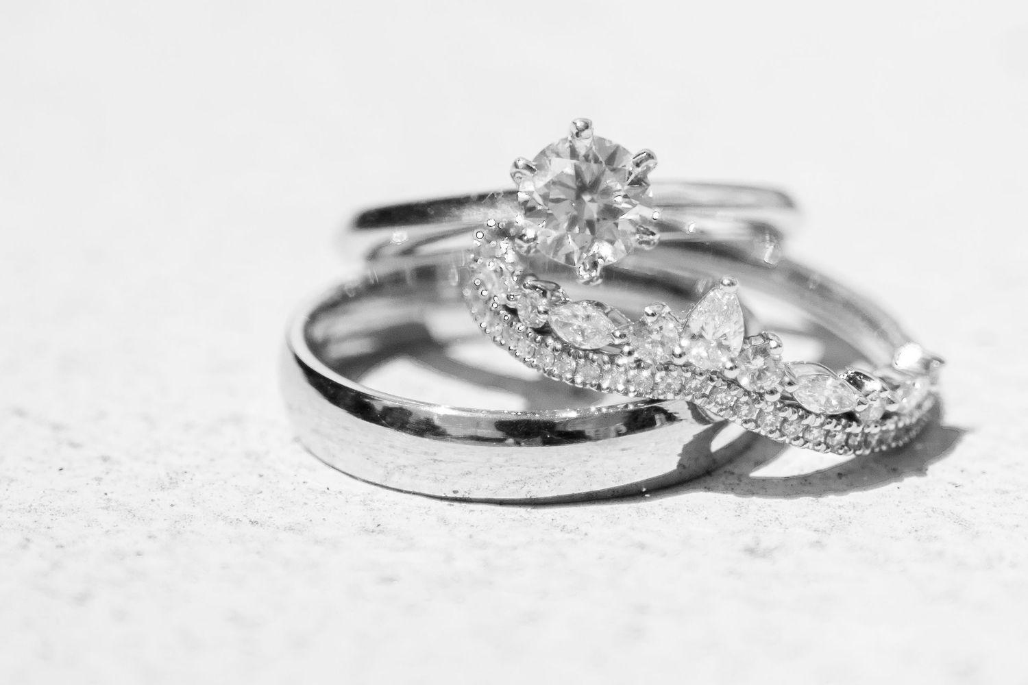 A close-up black and white photo of sparkling diamond engagement and wedding rings on a white textured surface.