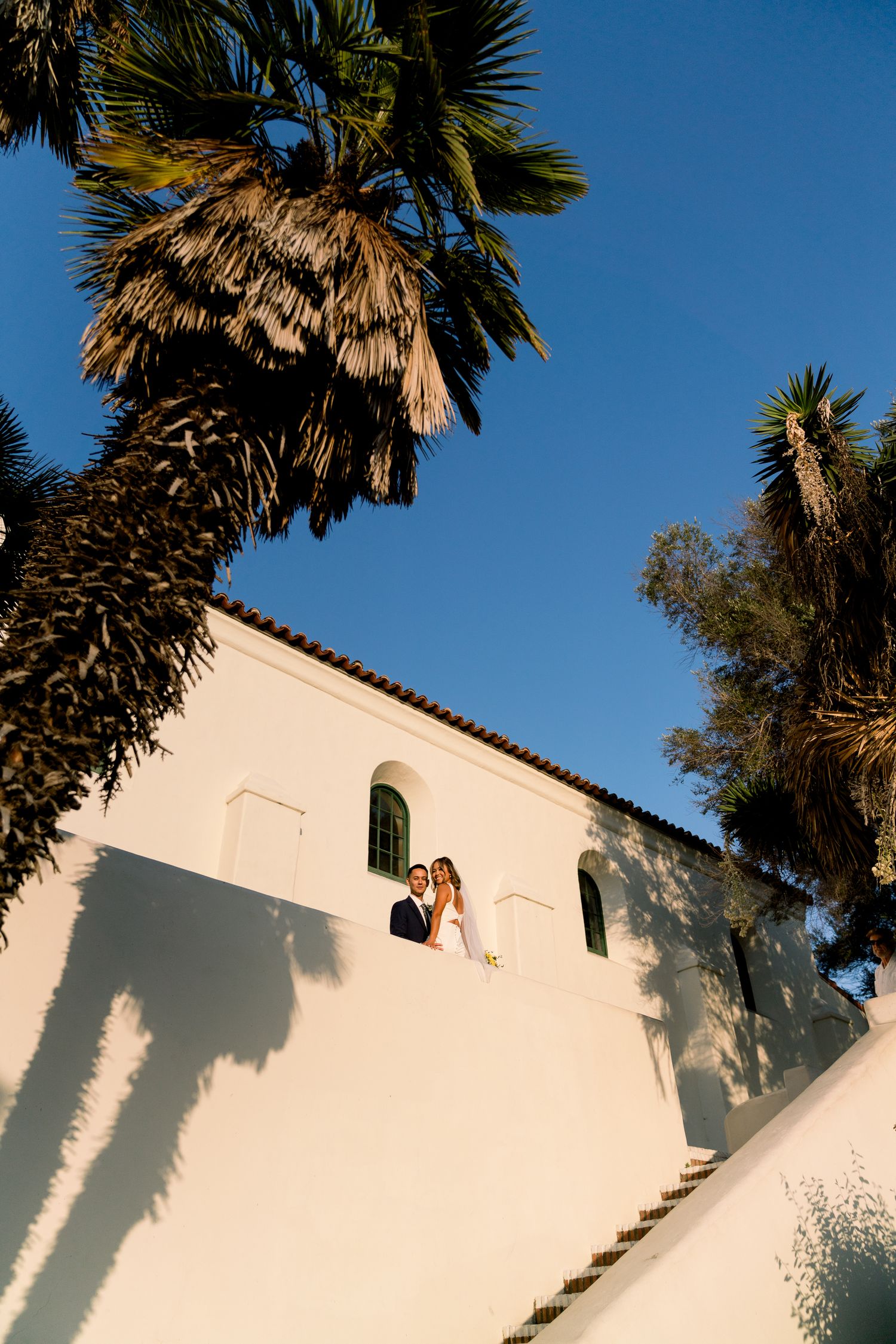 A couple stands on a white Spanish-style building balcony framed by palm trees against a bright blue sky.