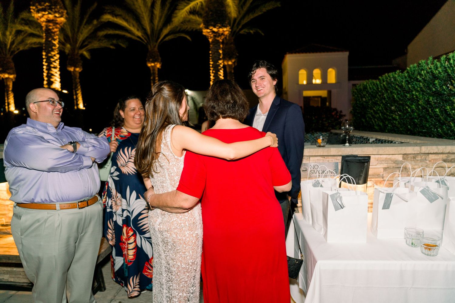 Group of party guests embrace and celebrate near illuminated palm trees at night.