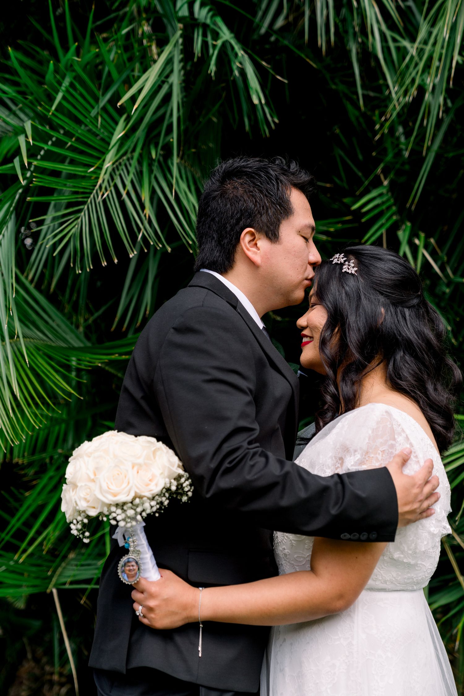 Bride and groom share a romantic embrace in front of lush tropical palm fronds during wedding photo session.