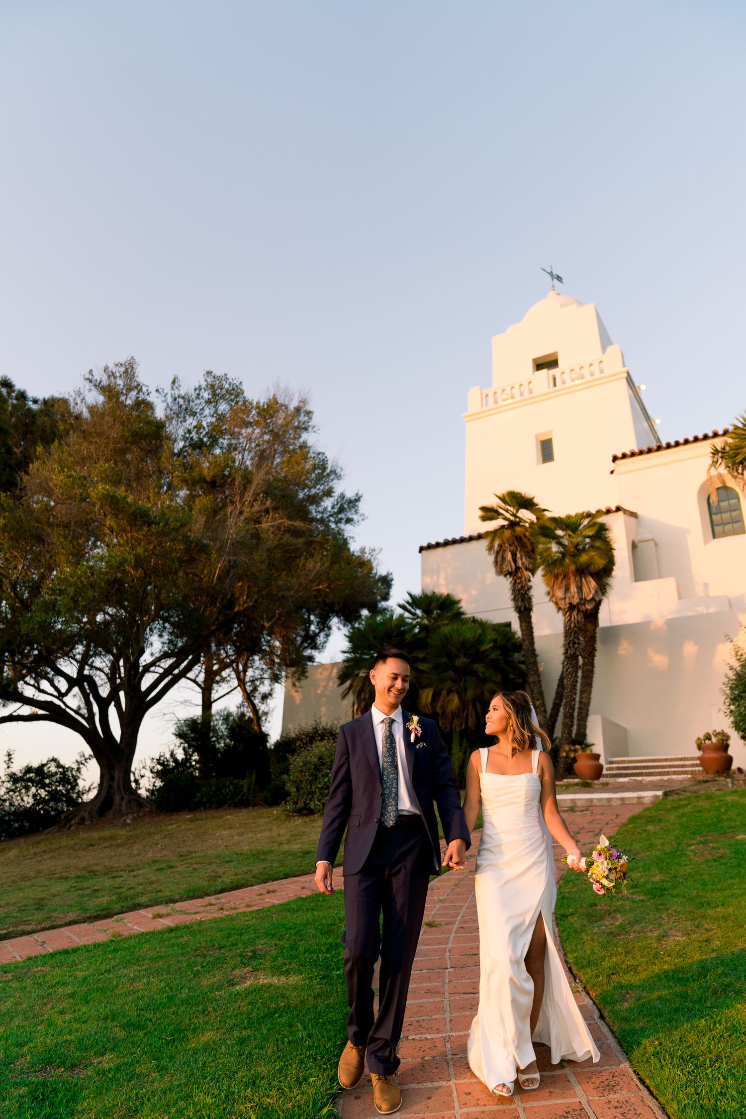 A couple walks down a path at sunset outside the iconic white Spanish-style La Jolla Presbyterian Church in San Diego.