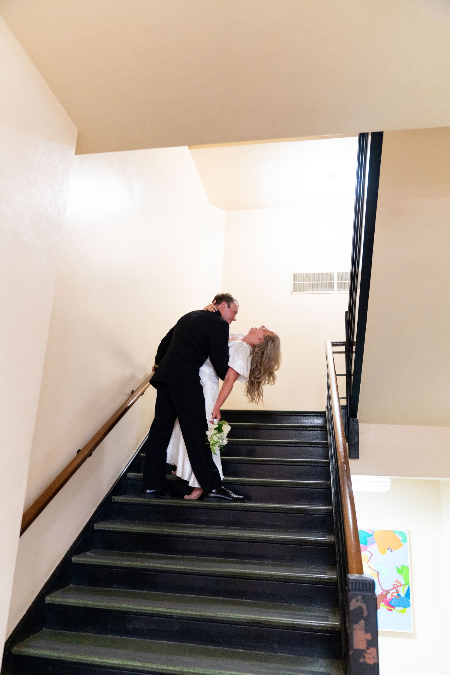 A couple in formal attire share a romantic moment on dark stairs in a light colored stairwell.