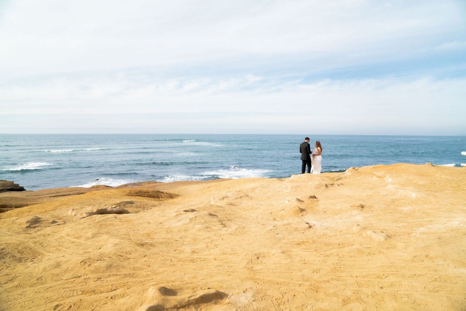 A couple stands on a coastal cliff overlooking the ocean during their wedding photoshoot.