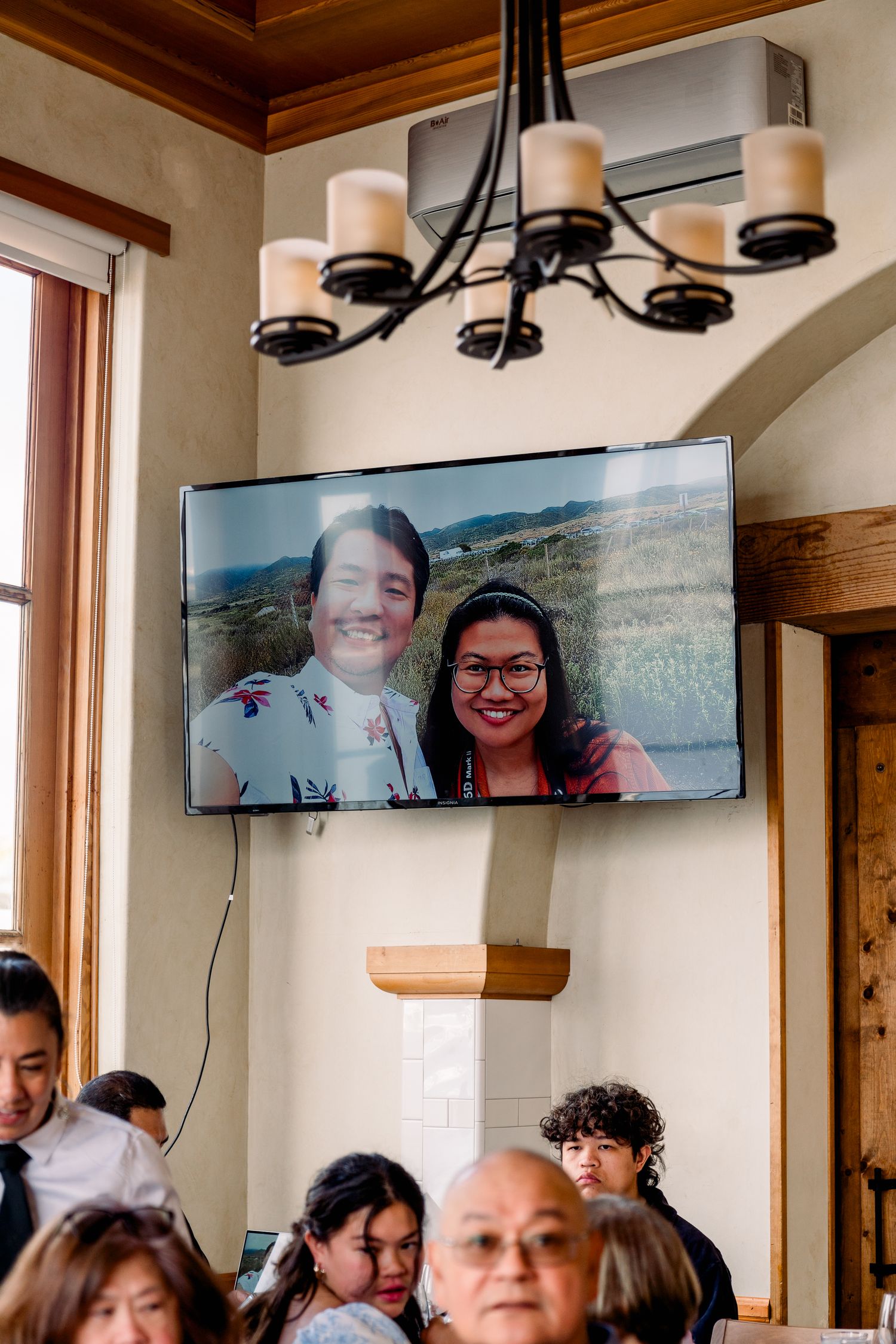 Wedding reception room with chandelier and video call displayed on wall-mounted TV.