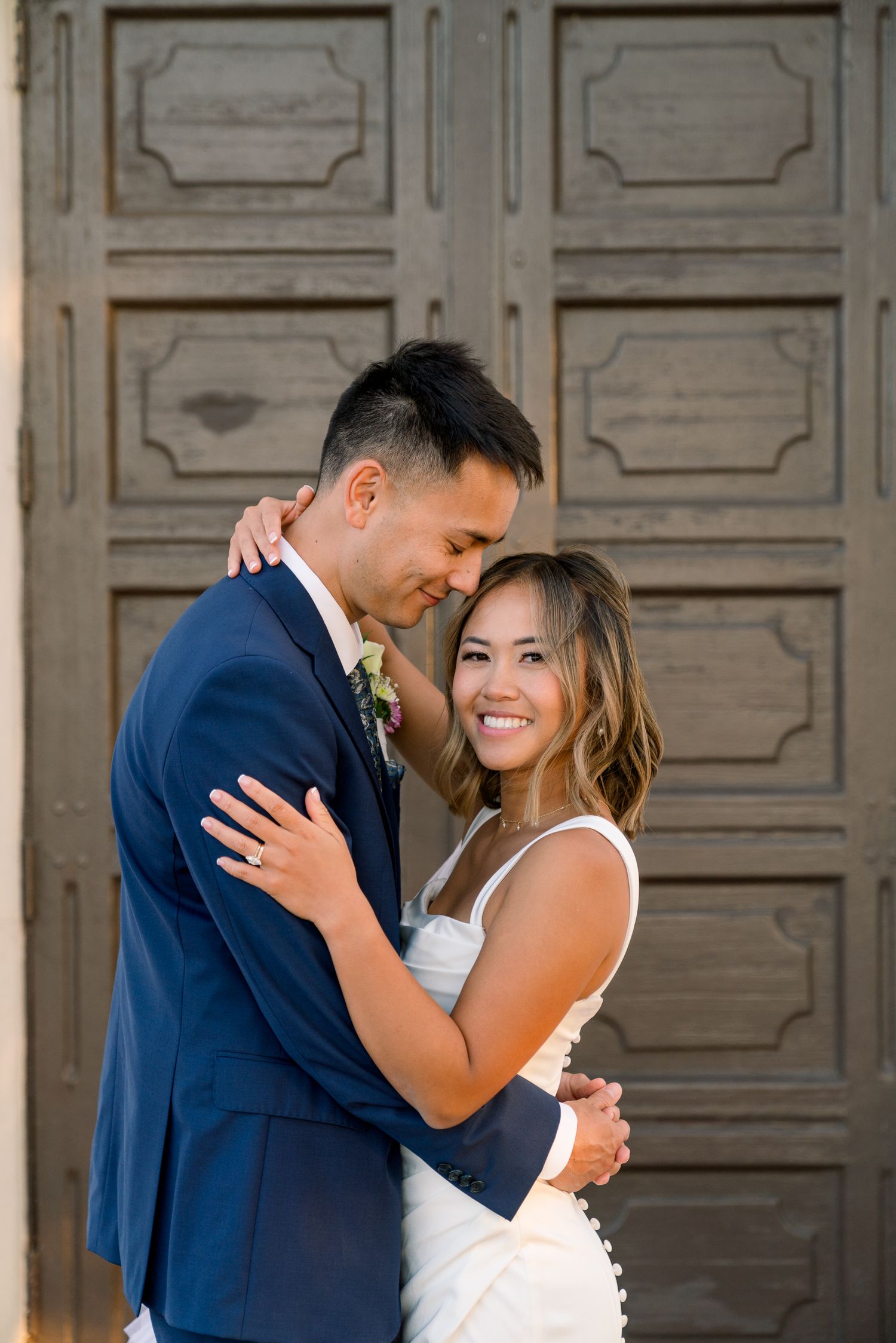 A couple embraces in front of ornate wooden doors during their wedding day photoshoot.