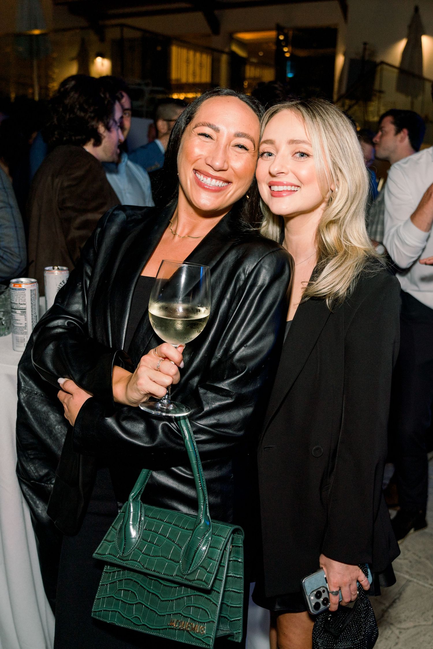 Two fashionable friends at a party, one with a green handbag and wine glass.