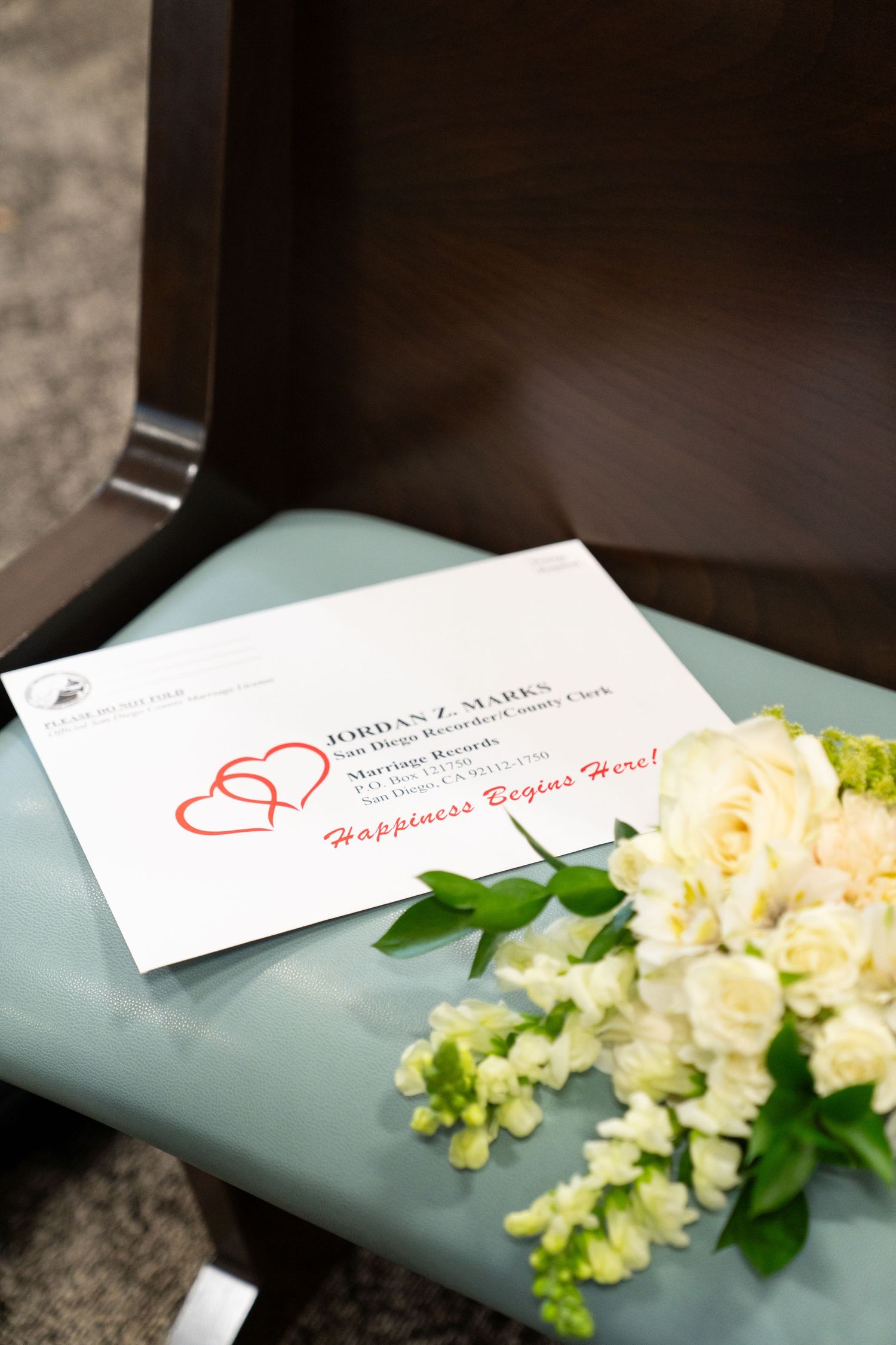 Wedding invitation card with red heart design lies beside cream colored roses and delicate white flowers on blue surface.